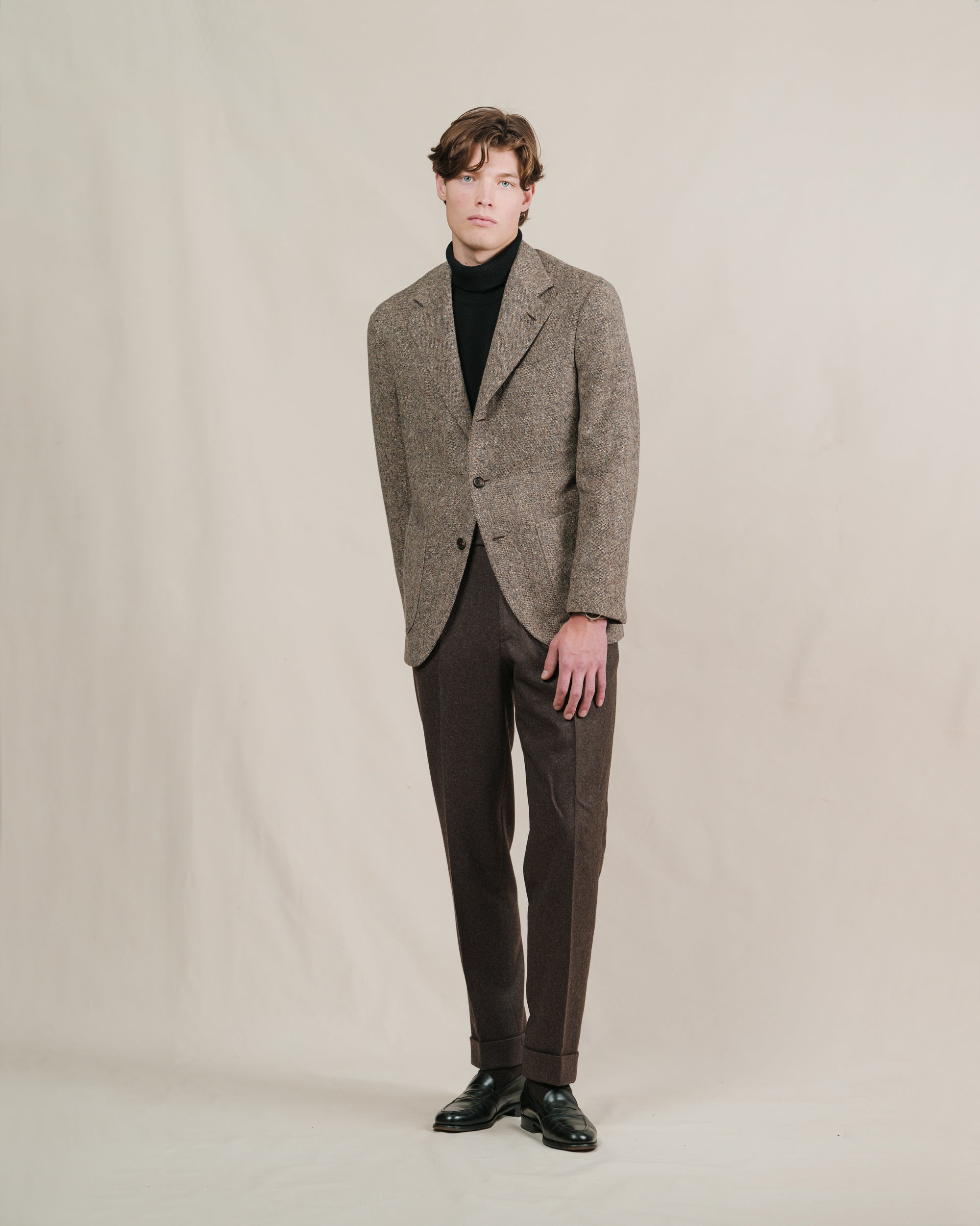 A man wearing a light brown donegal tweed sport coat, brown flannel trousers and a black roll neck