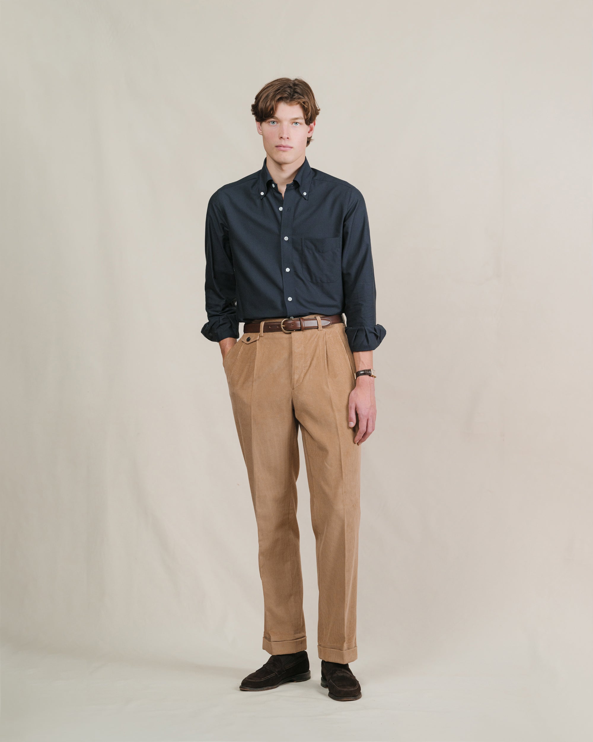Person wearing brown corduroy trousers, navy oxford cloth button down shirt, brown leather belt and suede penny loafers