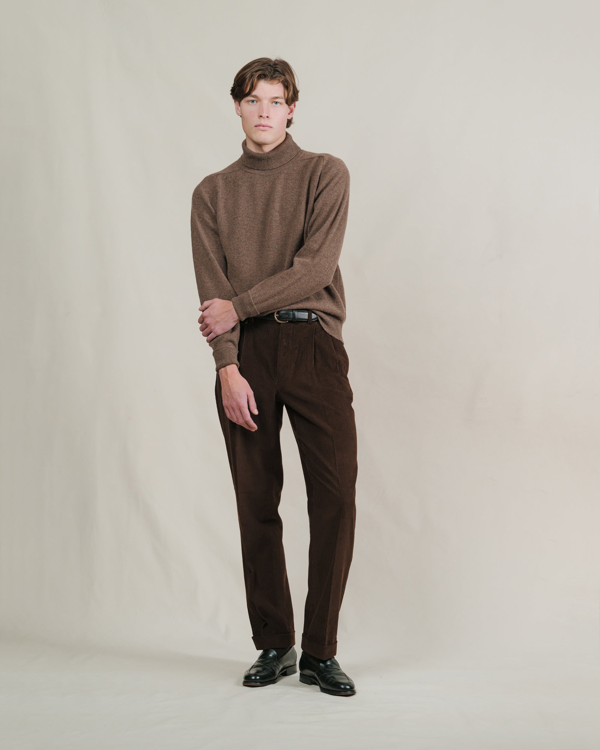 Man wearing a mid brown turtleneck sweater and corduroy pants against a plain background