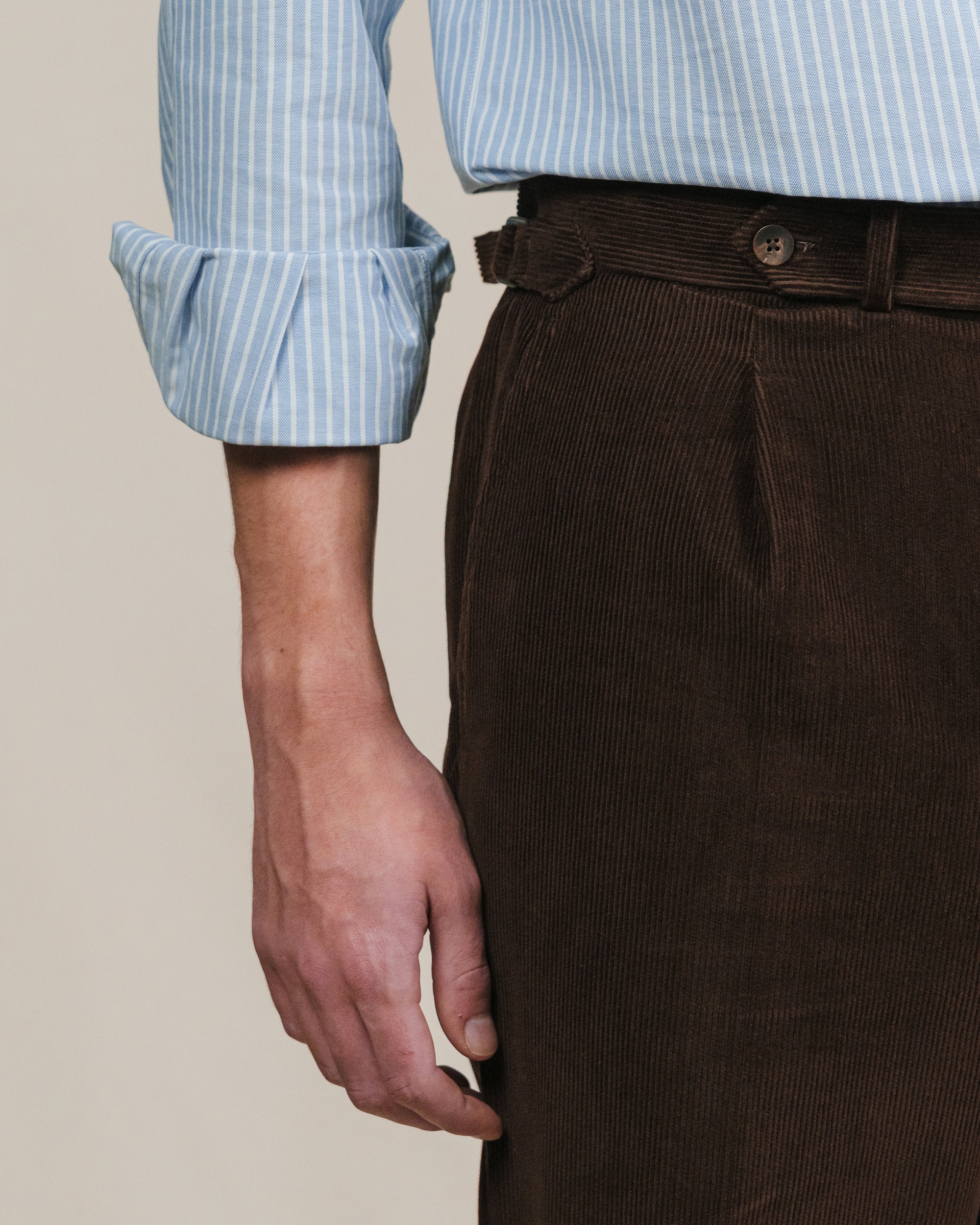 Brown corduroy pants with a light blue reverse striped oxford shirt on a beige background