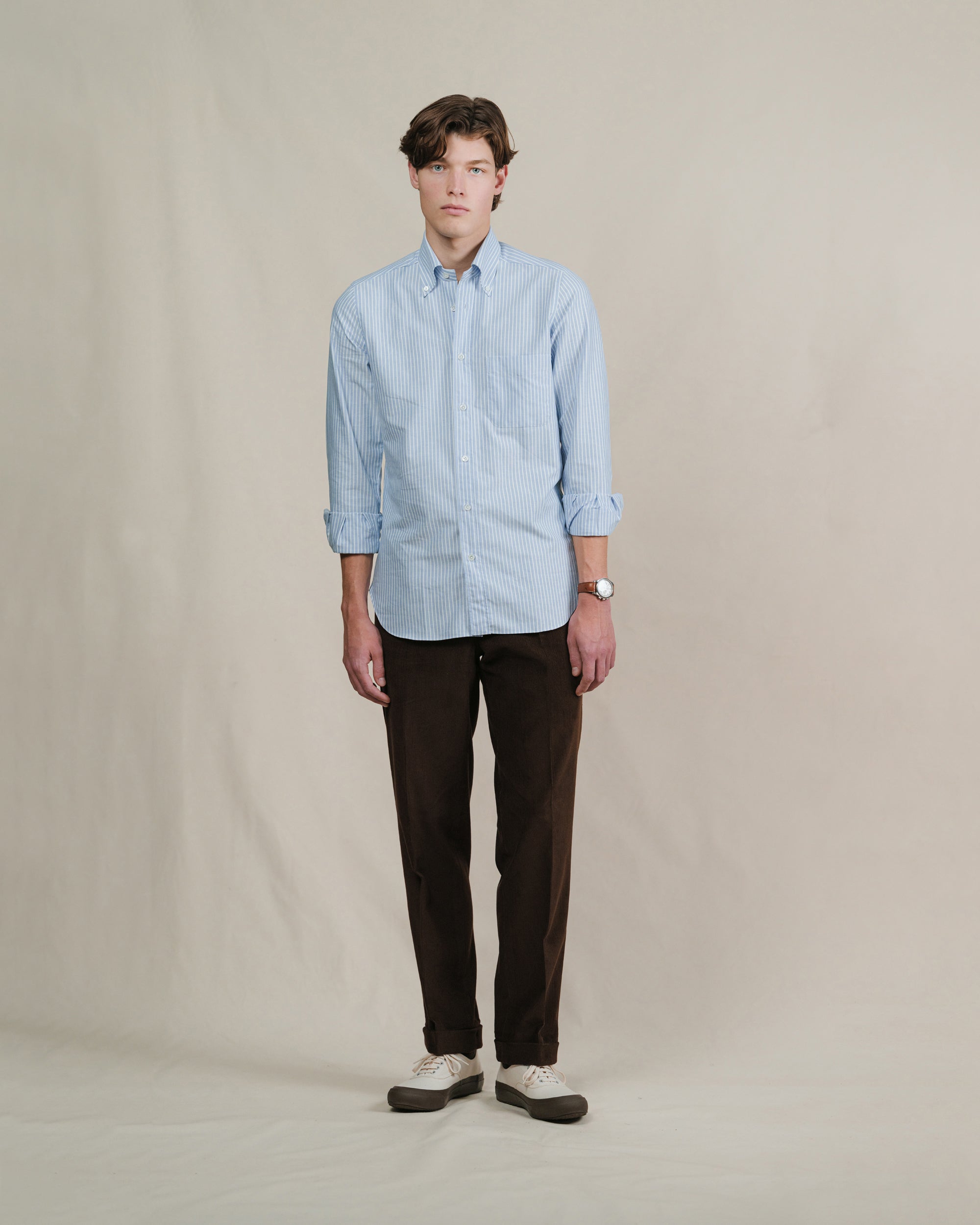 Man wearing a blue reverse stripe oxford cloth button down shirt and dark brown corduroy pants on a beige background