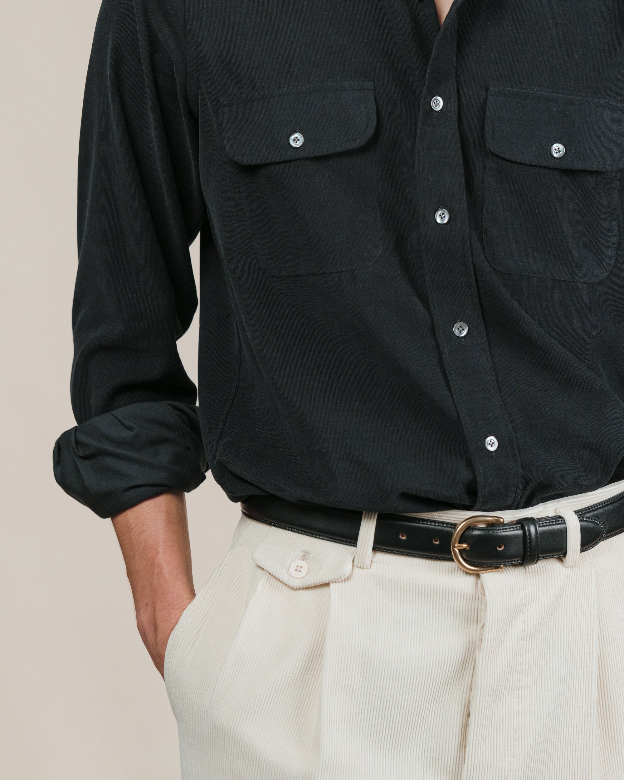 Person wearing a black shirt with two chest pockets, cream corduroy trousers and a black dress belt with brass buckle
