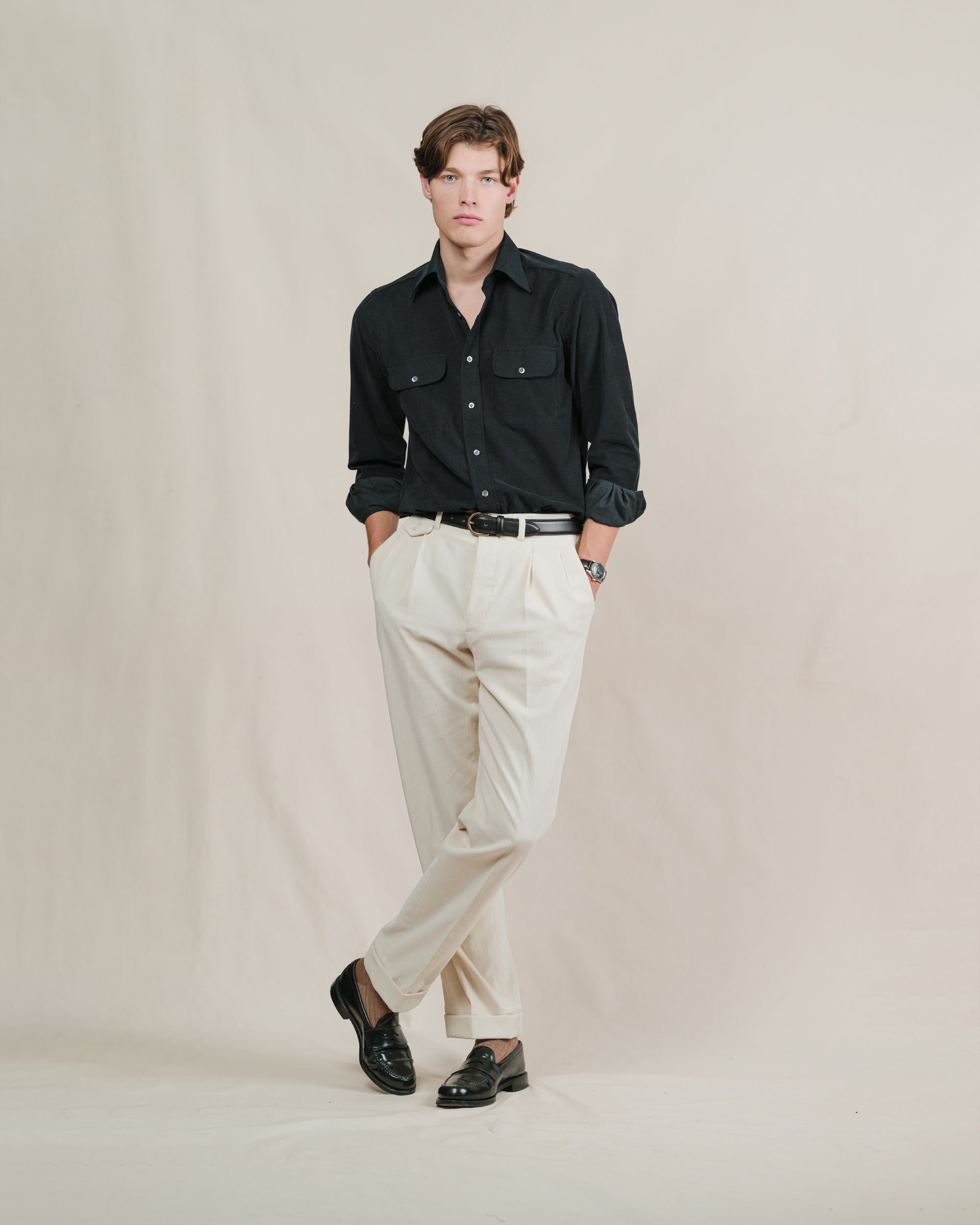 Man wearing a black corduroy shirt, cream pants, black belt and black penny loafers against a plain background