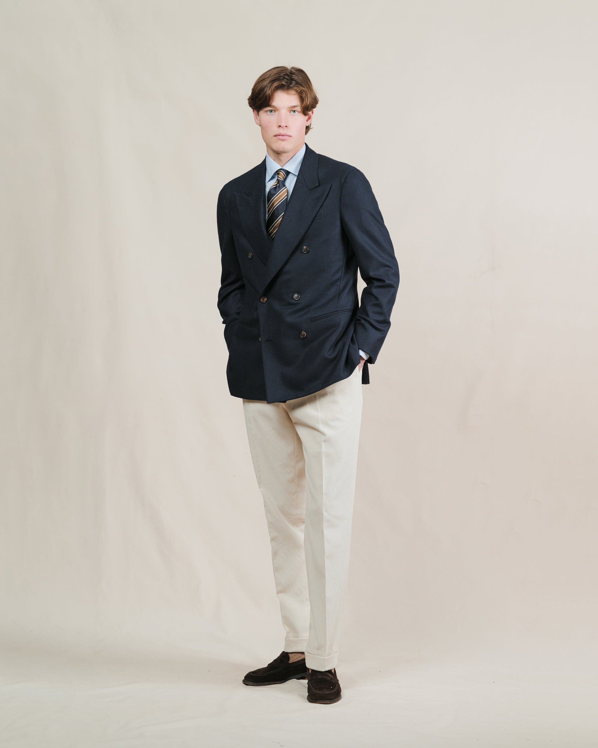 Man wearing a navy double-breasted jacket, cream corduroy pants, and a striped tie on a plain background