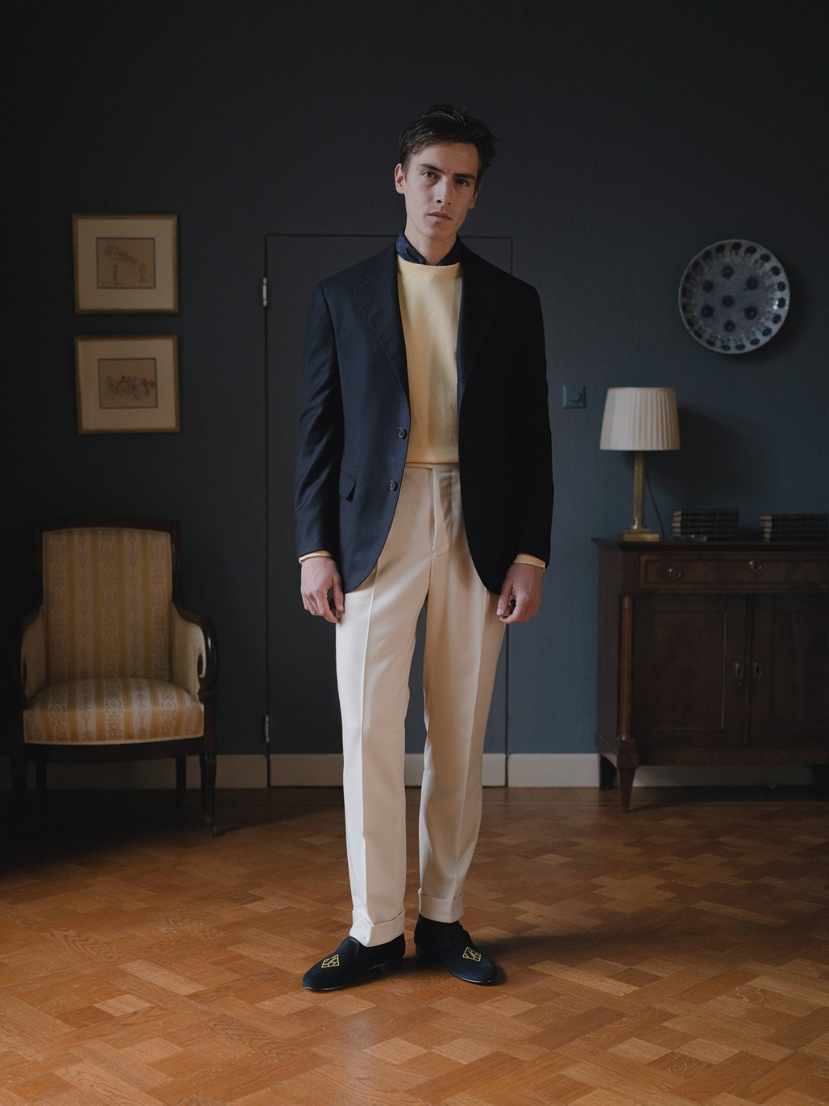 Man wearing a navy wool cashmere sport coat, a yellow crewneck sweater, cream flannels and prince albert slippers