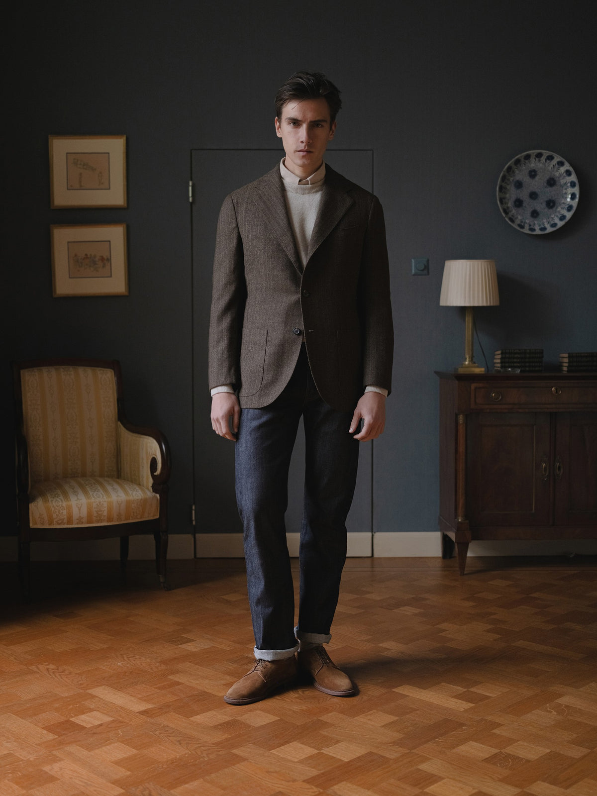 Man wearing a brown herringbone sport coat, raw denim jeans, light brown crewneck sweater and snuff suede cap toe bluchers