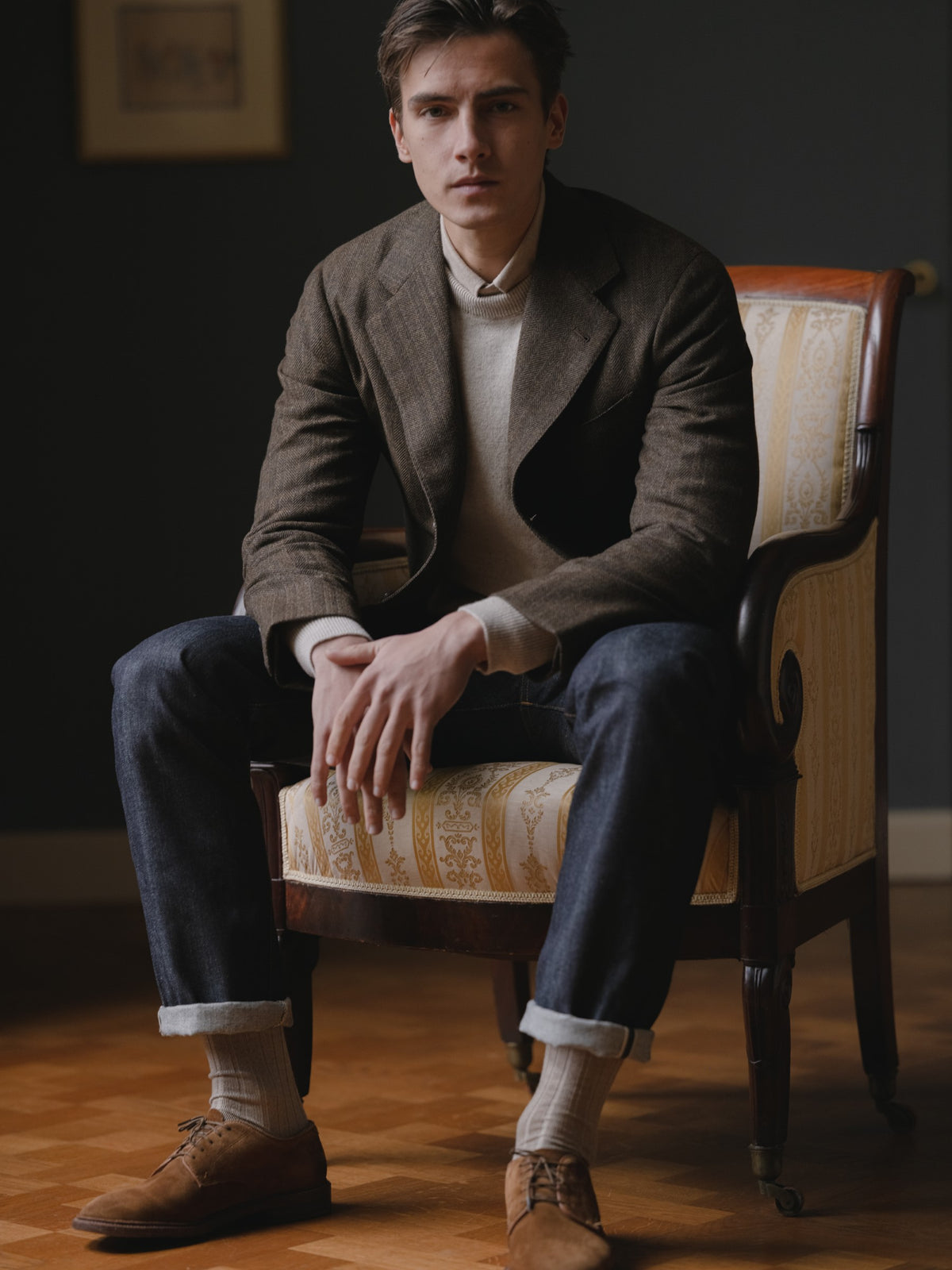 Man wearing a brown herringbone sport coat, raw denim jeans, light brown crewneck sweater and snuff suede cap toe bluchers