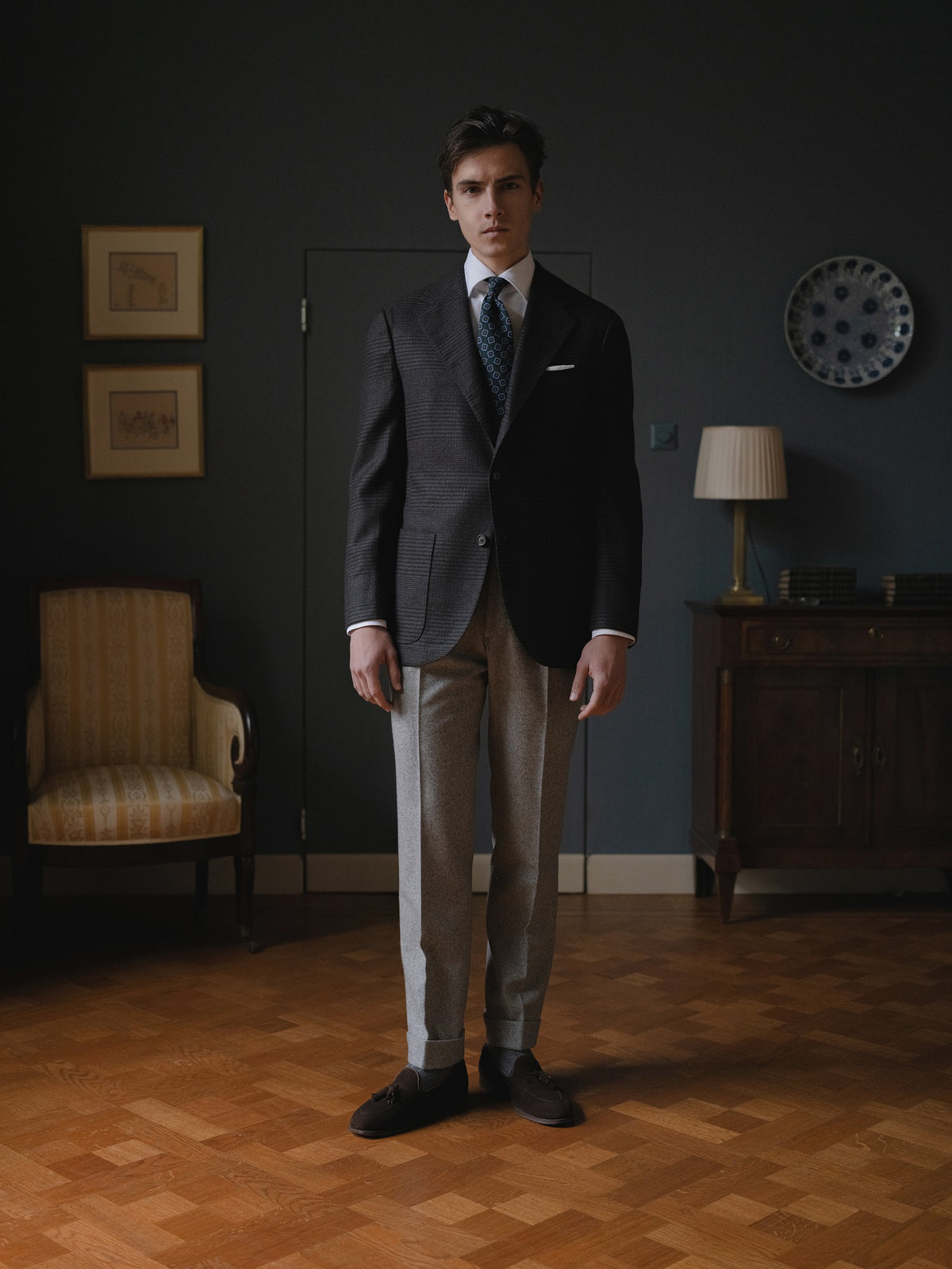 Man wearing a dark grey prince of wales sport coat, light grey flannel trousers, white shirt and green medallion ancient madder tie