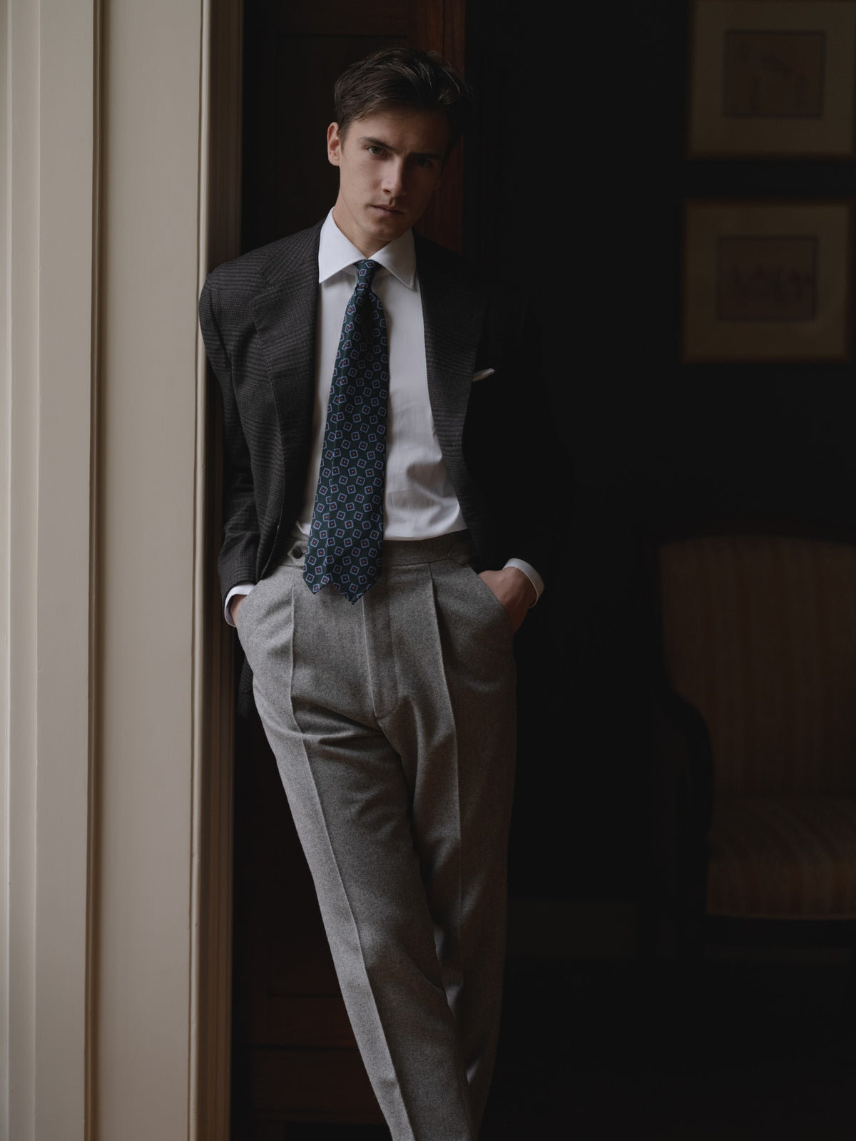 Man wearing a dark grey prince of wales sport coat, light grey flannel trousers, white shirt and green medallion ancient madder tie