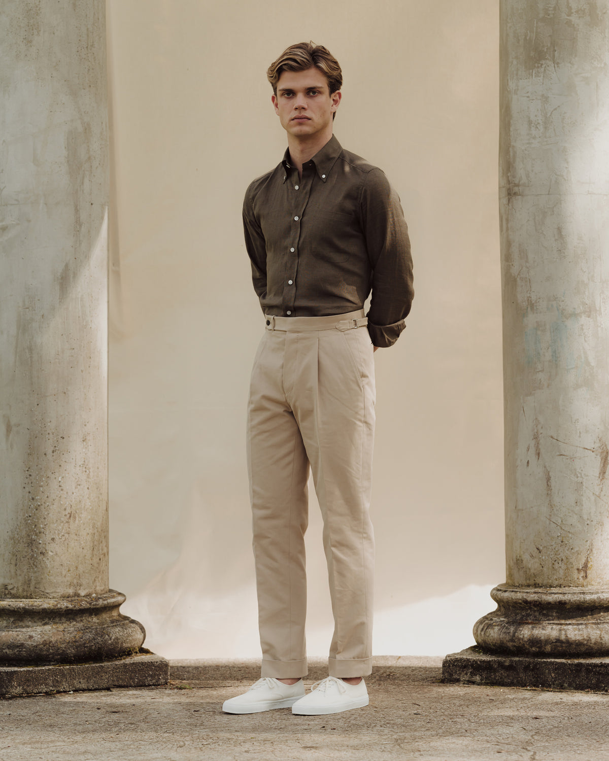 Man wearing stone cotton drill trousers, olive green linen button down shirt and white tennis sneakers
