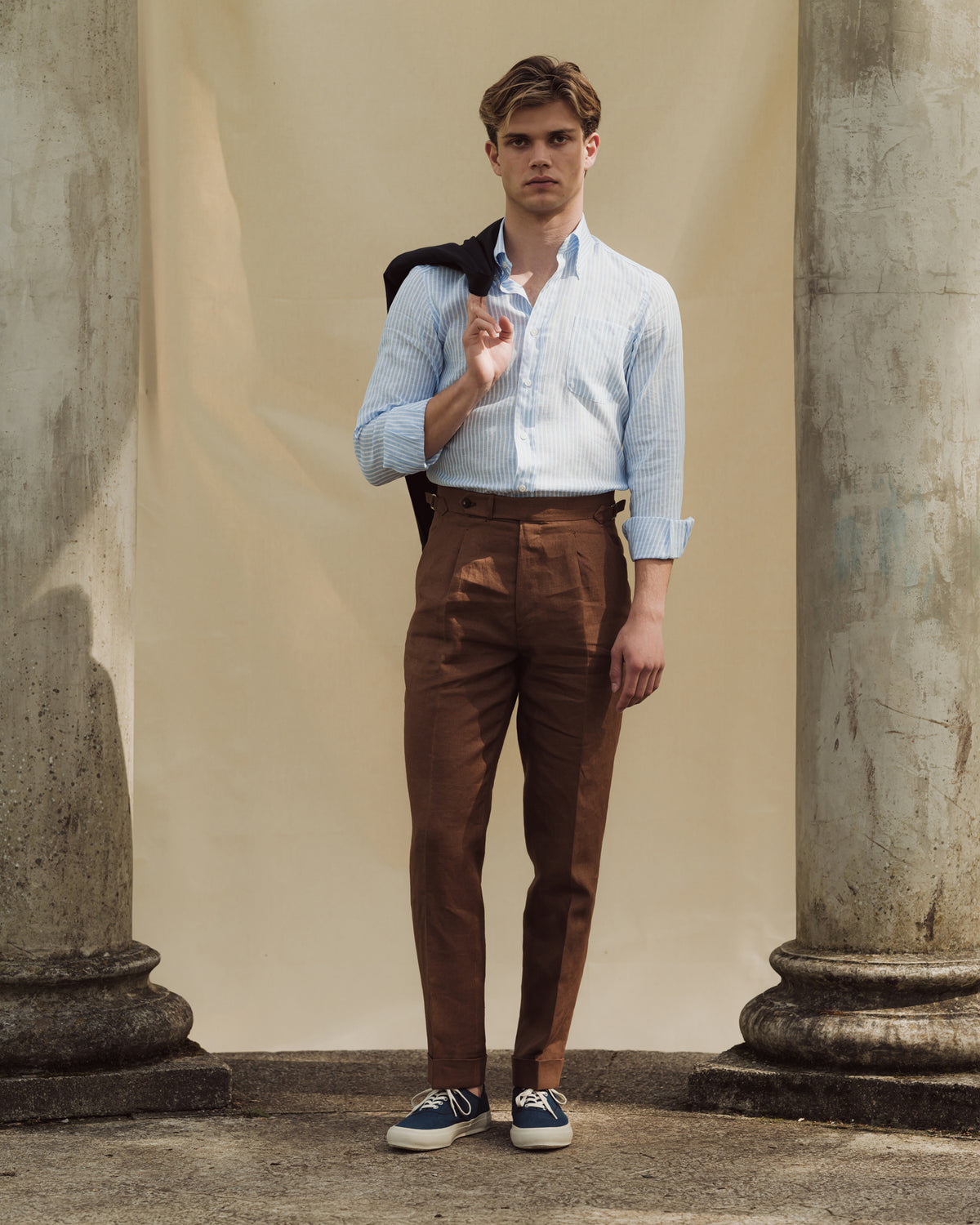 Man wearing dark tan single pleat trousers, striped linen button down shirt and dark blue canvas deck shoes