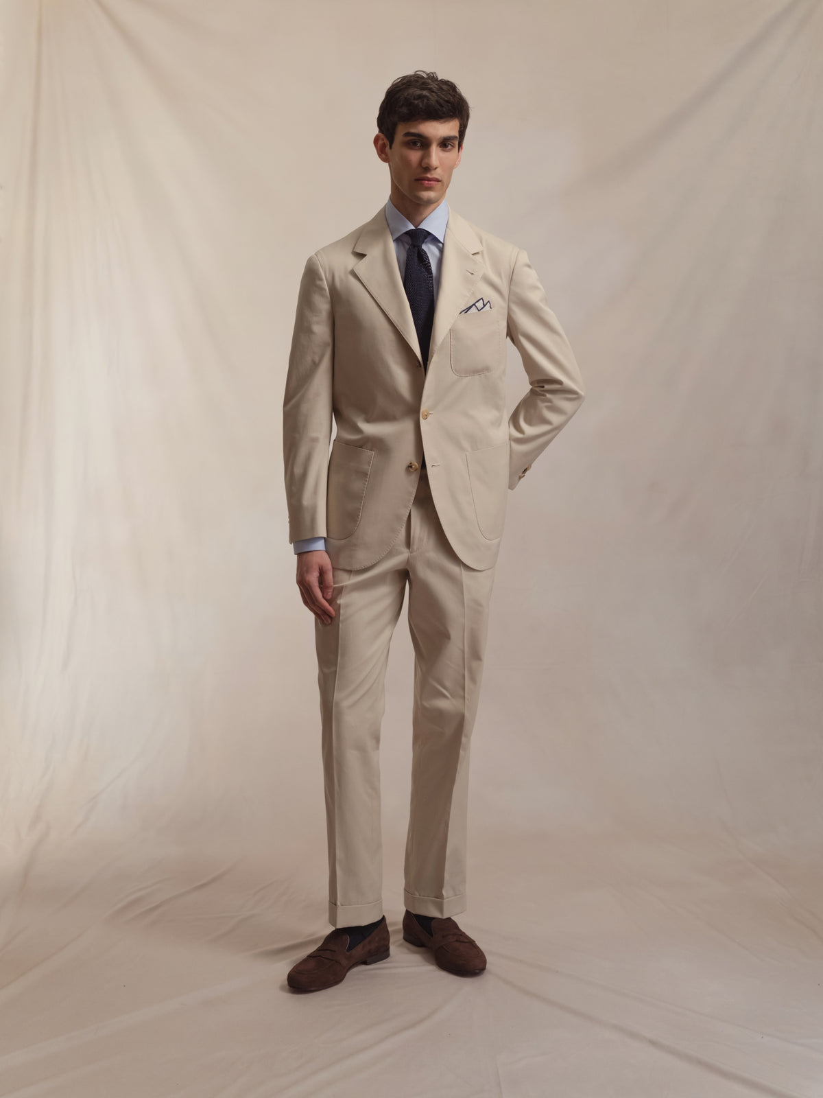 Man wearing a beige cotton suit with a navy knitted tie against a plain background