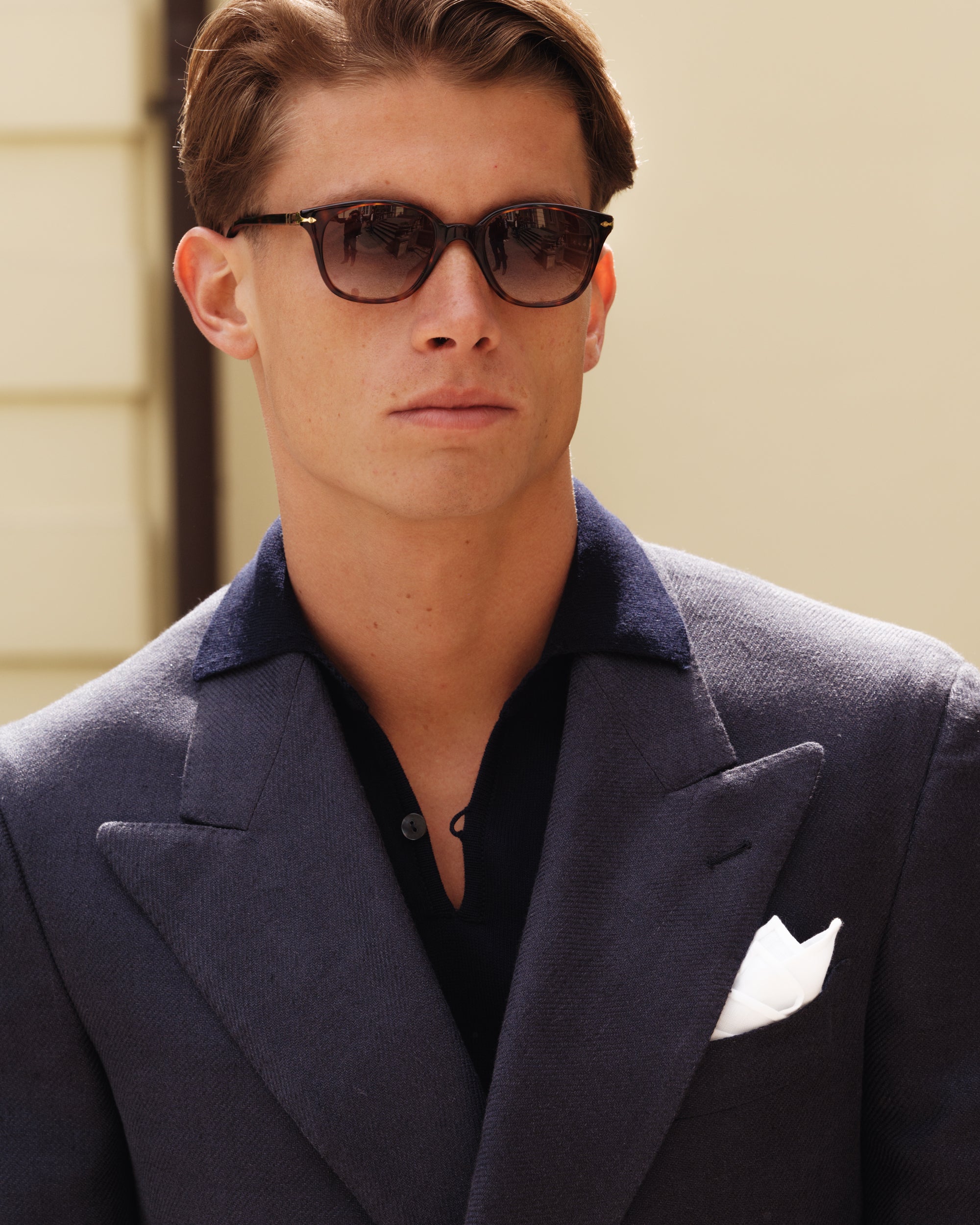 Close-up of a man wearing a navy double-breasted jacket with wide peak lapels, navy knit polo, white pocket square, and tortoiseshell sunglasses — refined spring-summer menswear look by Besnard.