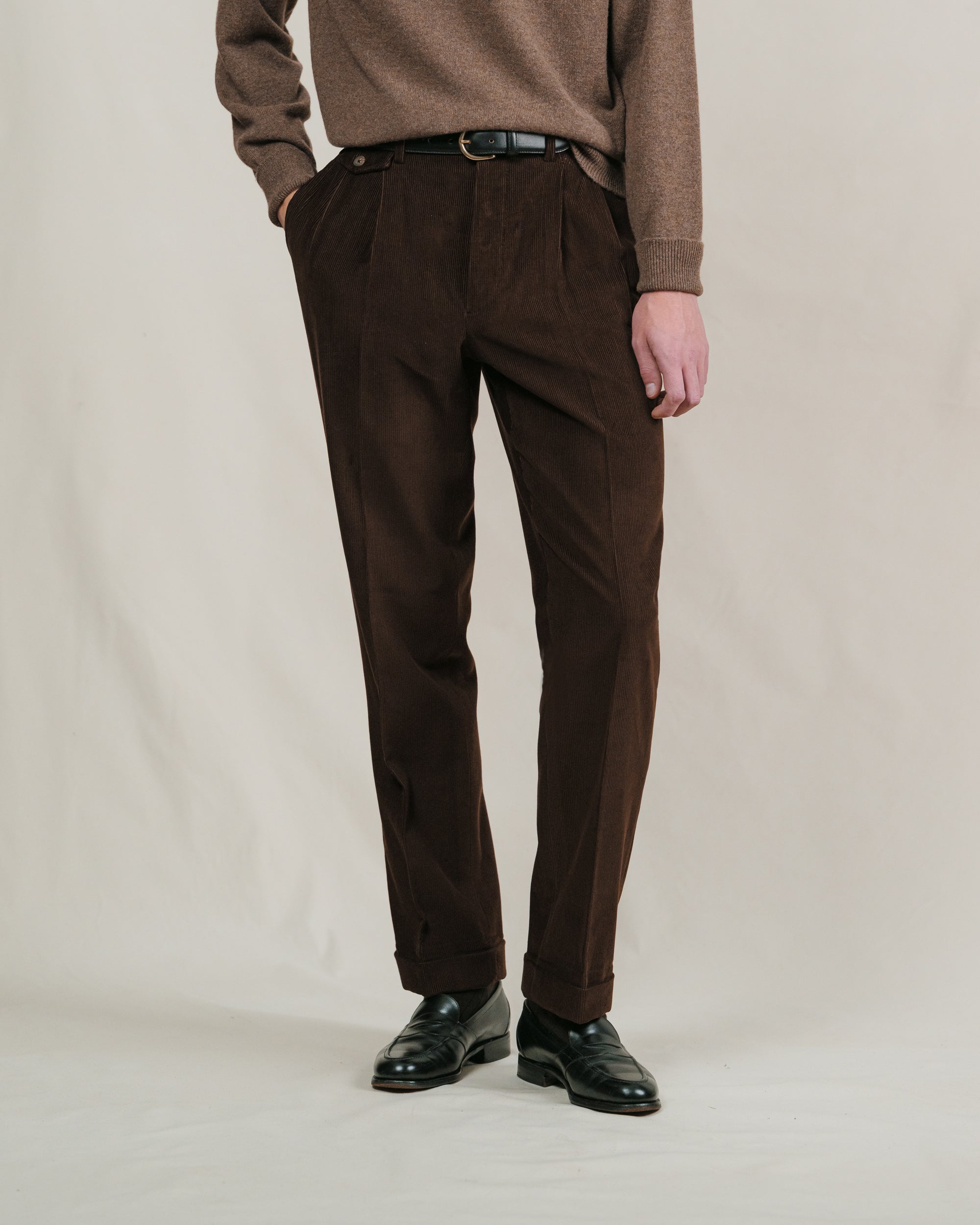 Person wearing brown corduroy trousers, a black leather belt and a brown sweater on a plain background