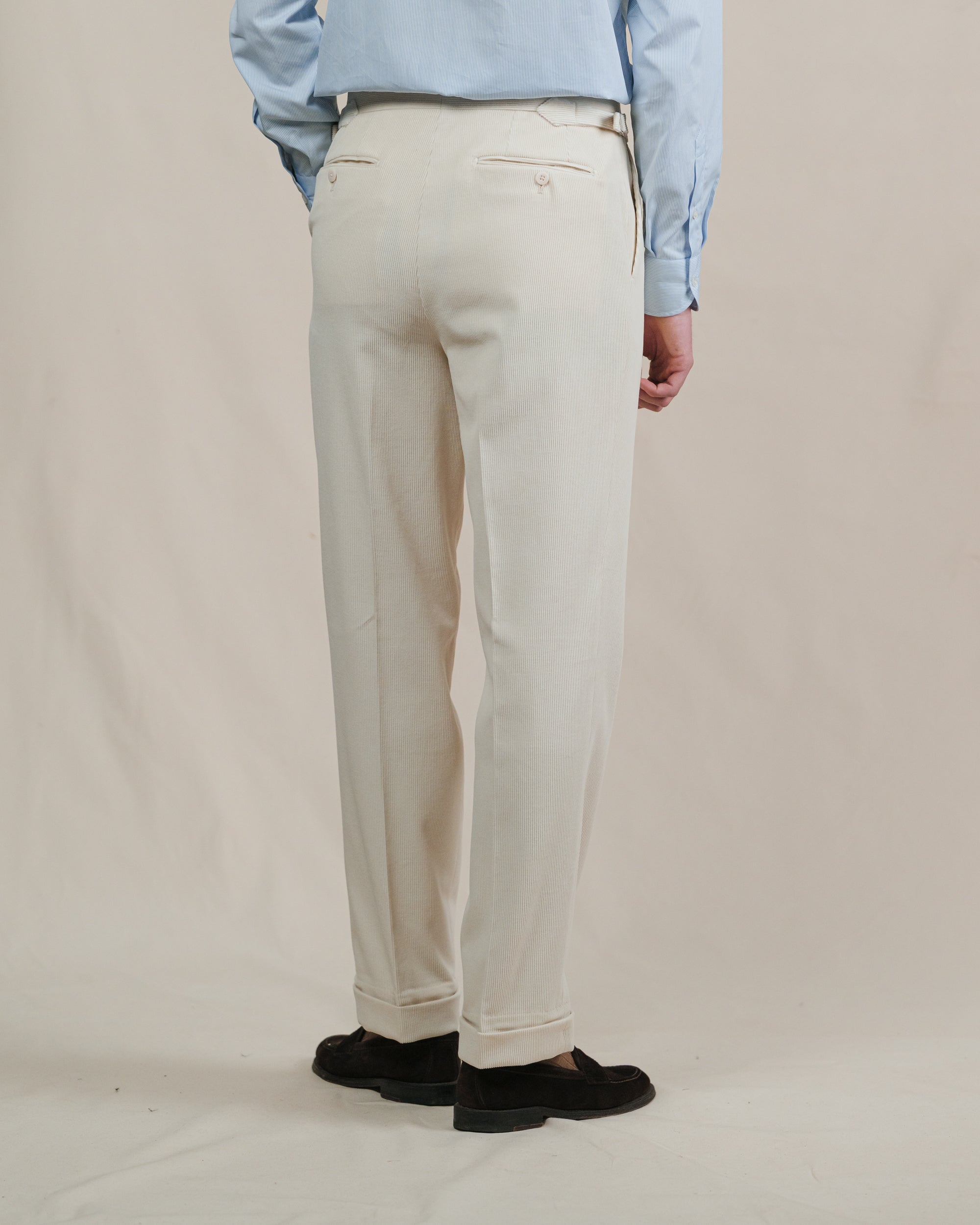 Person wearing cream corduroy trousers, brown suede penny loafers and a light blue shirt on a plain background