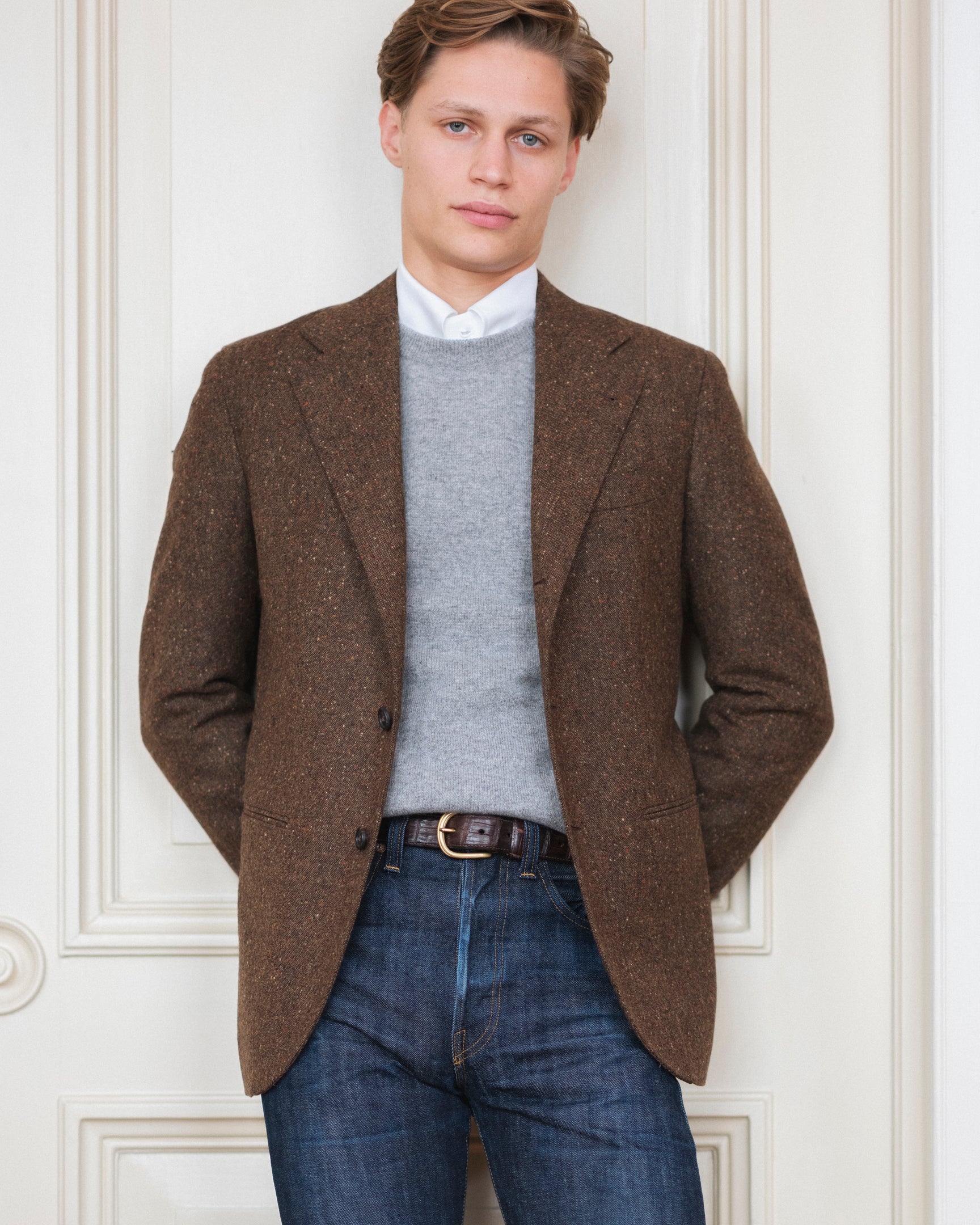 Man wearing a pair of raw denim jeans, white shirt, light grey crewneck sweater and a brown Donegal tweed sport coat