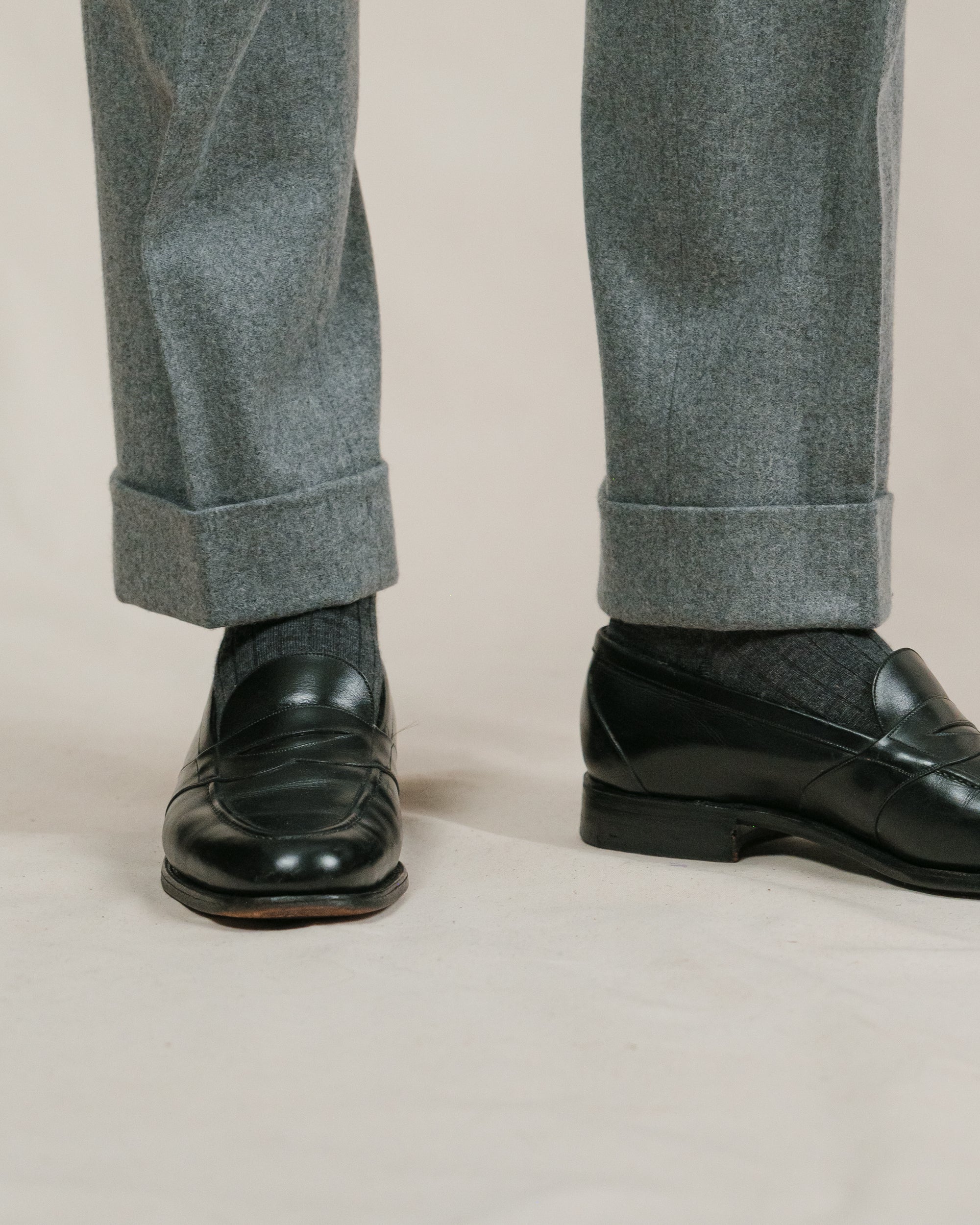 Man wearing a pair of light grey flannel trousers, dark grey socks and black leather full strap penny loafers