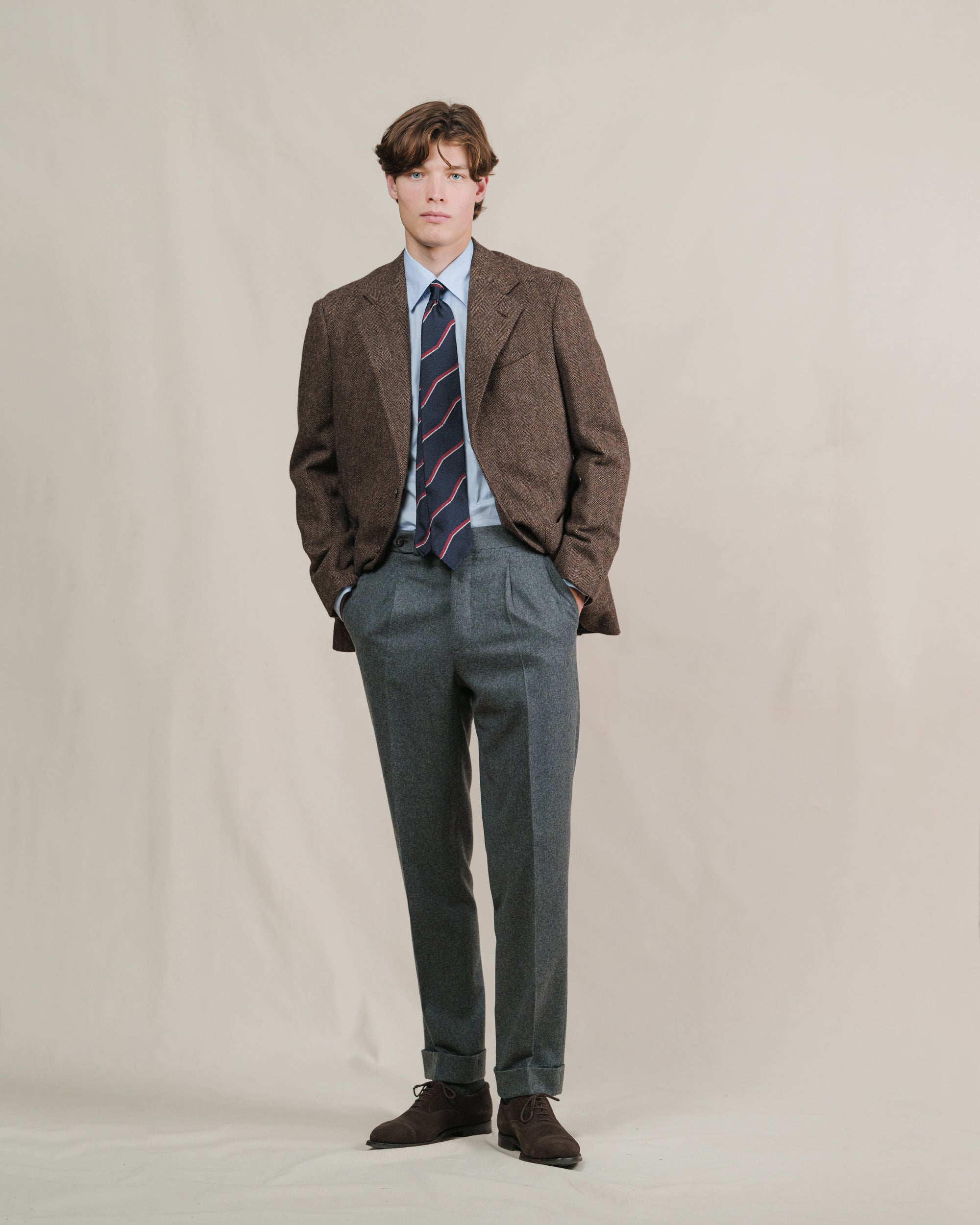 A person wearing a brown herringbone tweed sport coat, a pair of mid grey flannel trousers, a light blue shirt and a repp tie
