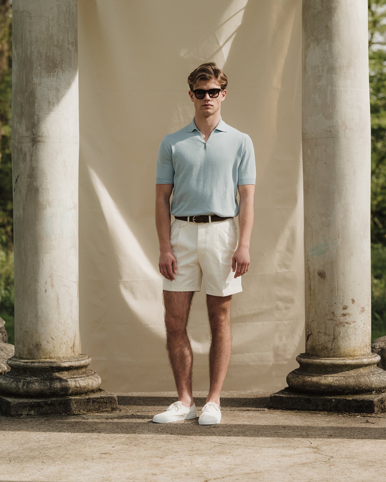 Man wearing a light blue cotton skipper polo, white shorts, brown suede belt and white tennis sneakers