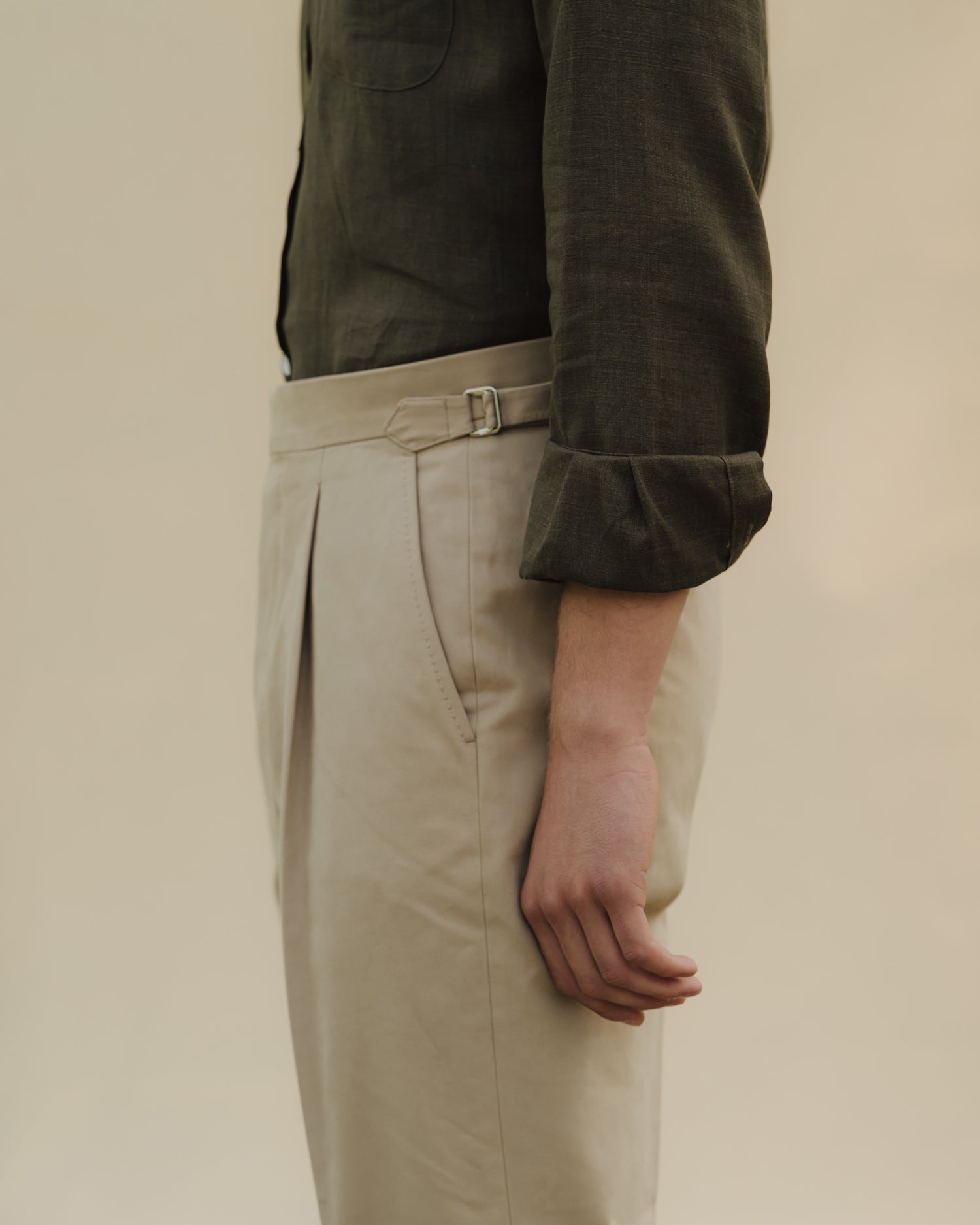 Man wearing stone cotton drill trousers with single pleats and side adjusters combined with an olive green linen button down shirt