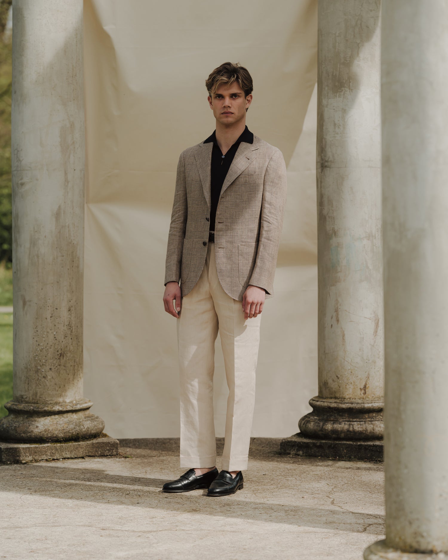 Man wearing stone linen trousers, navy cotton skipper polo and beige brown linen prince of wales sport coat