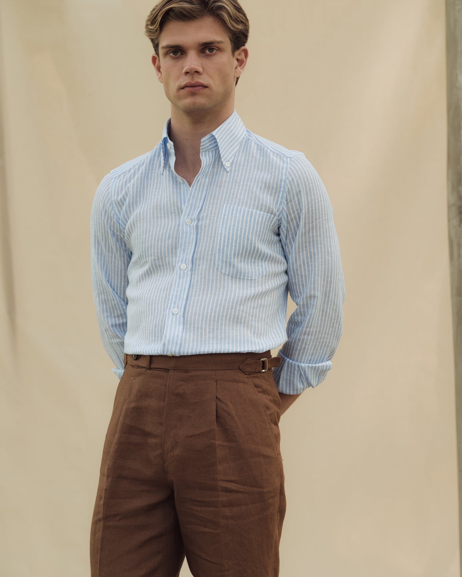 Man wearing dark tan trousers with single pleats and side adjusters, and a reverse striped linen button down shirt