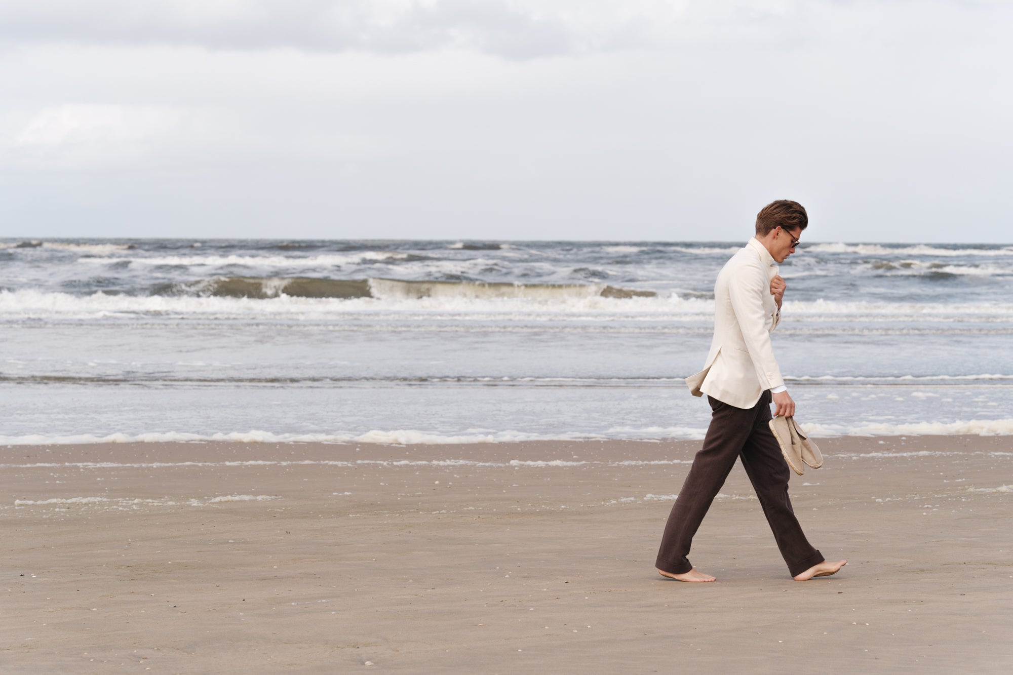 Man in cream jacket and brown linen trousers walking barefoot along a windy beach, holding espadrilles in hand — spring-summer menswear look by Besnard