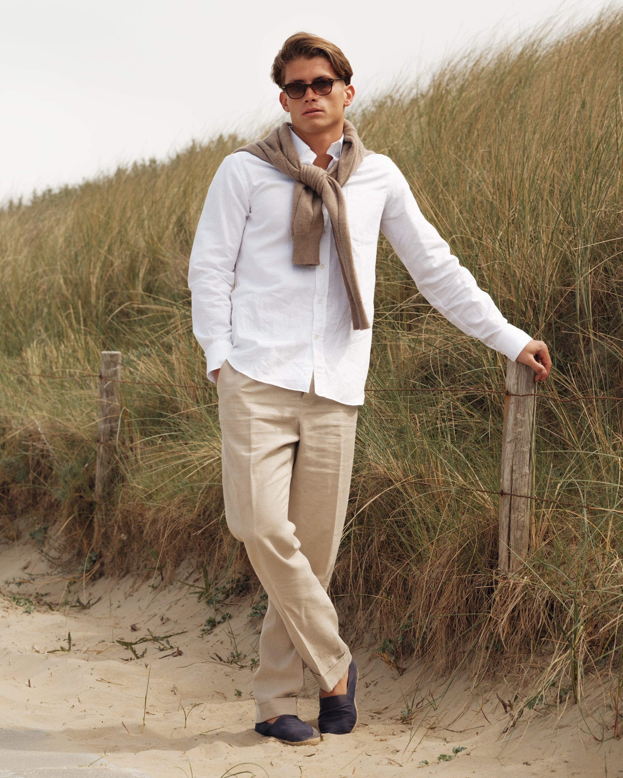Man wearing a white cotton-linen button-down shirt, natural linen trousers, and navy espadrilles with a light brown cashmere-cotton jumper draped over his shoulders — relaxed spring-summer menswear look by Besnard, photographed in coastal dunes.