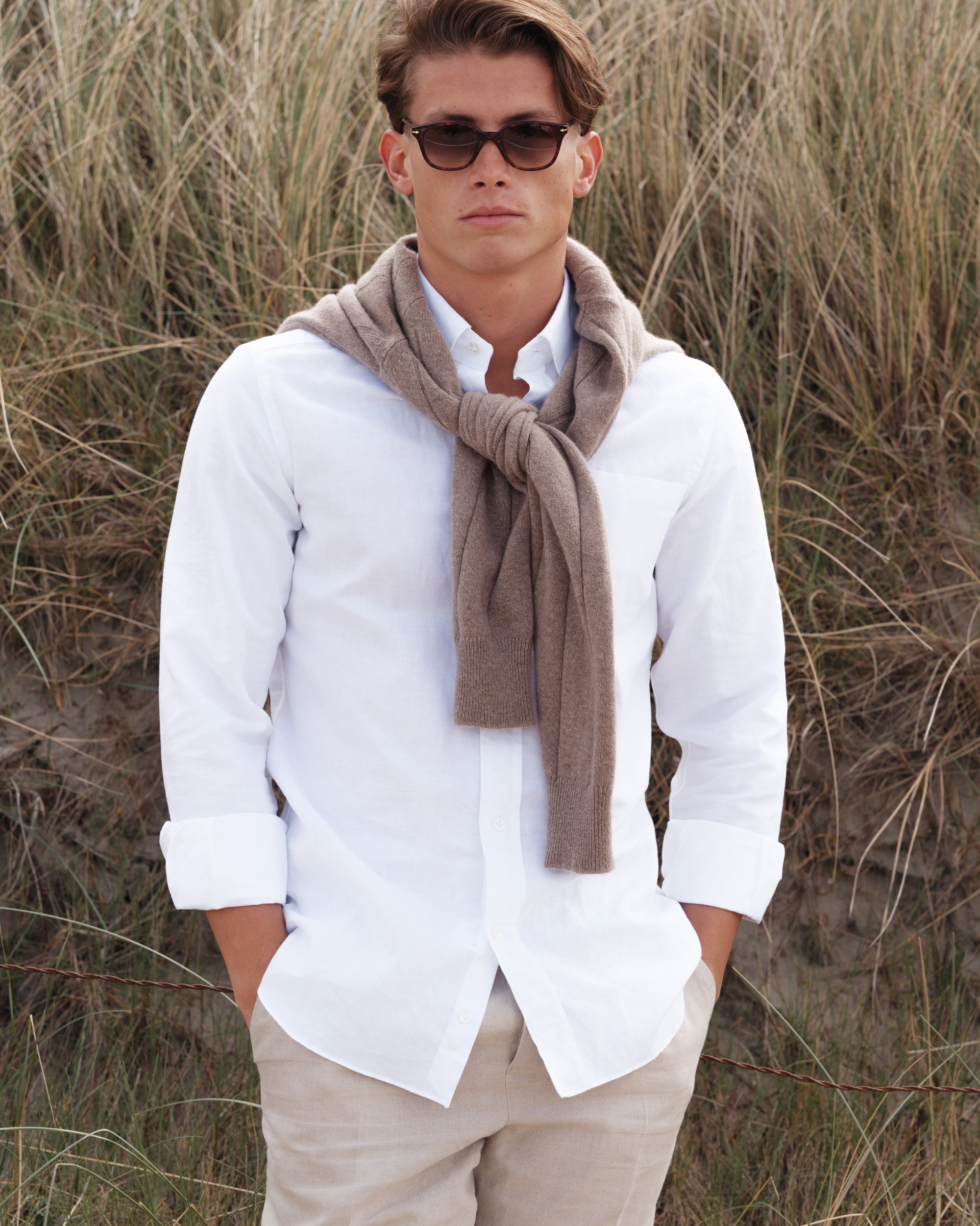 Man wearing a white cotton-linen button-down shirt and natural linen trousers with a light brown cashmere-cotton jumper draped over his shoulders — relaxed spring-summer menswear look by Besnard, photographed in coastal dunes.