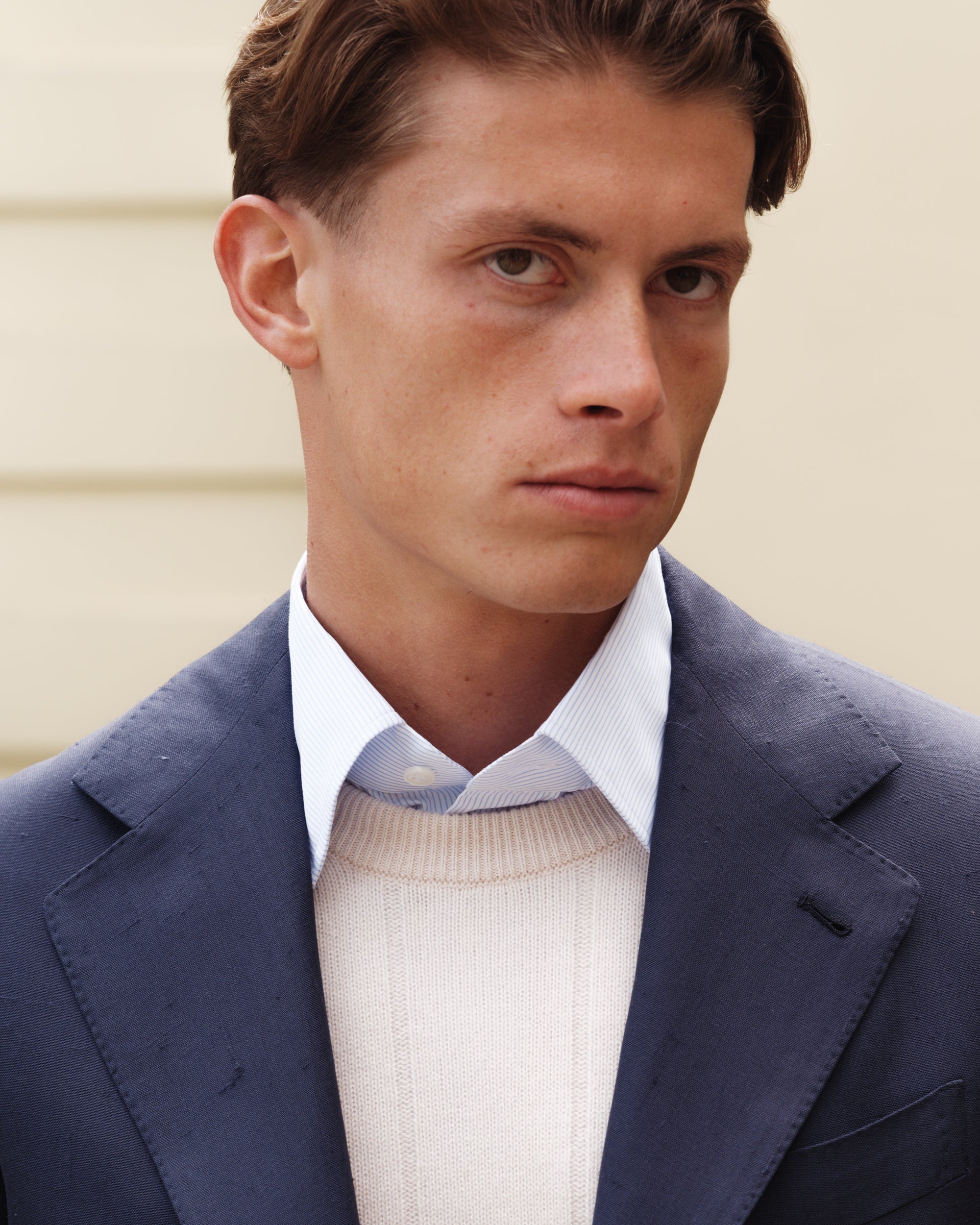 Close-up of a man wearing a navy silk jacket, cream cashmere-cotton crew-neck jumper, and blue striped shirt — refined spring-summer layering by Besnard.