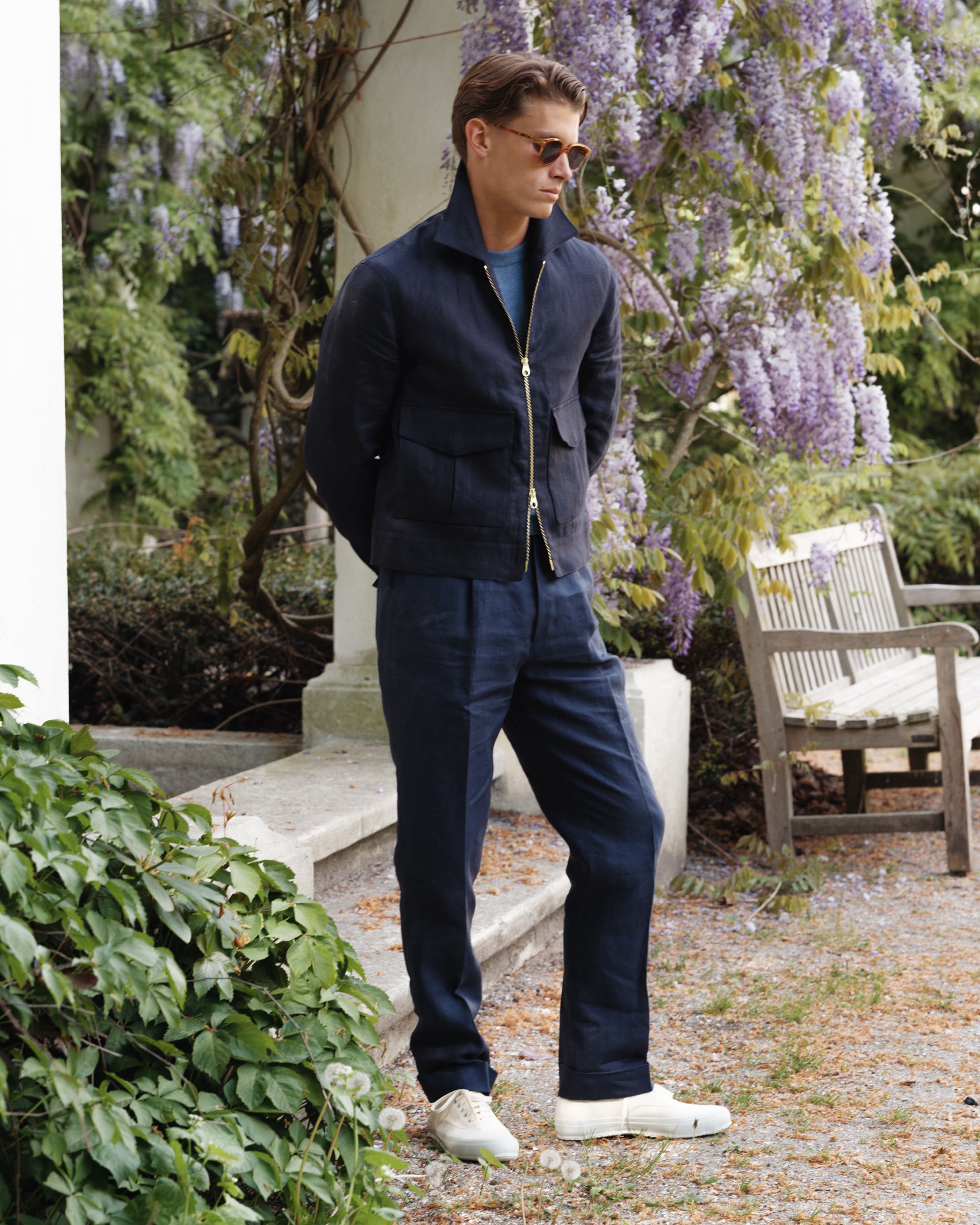 Man wearing a navy linen flight jacket and matching trousers with a blue crew-neck shirt and white tennis sneakers, standing under wisteria in a garden — spring-summer menswear look by Besnard.