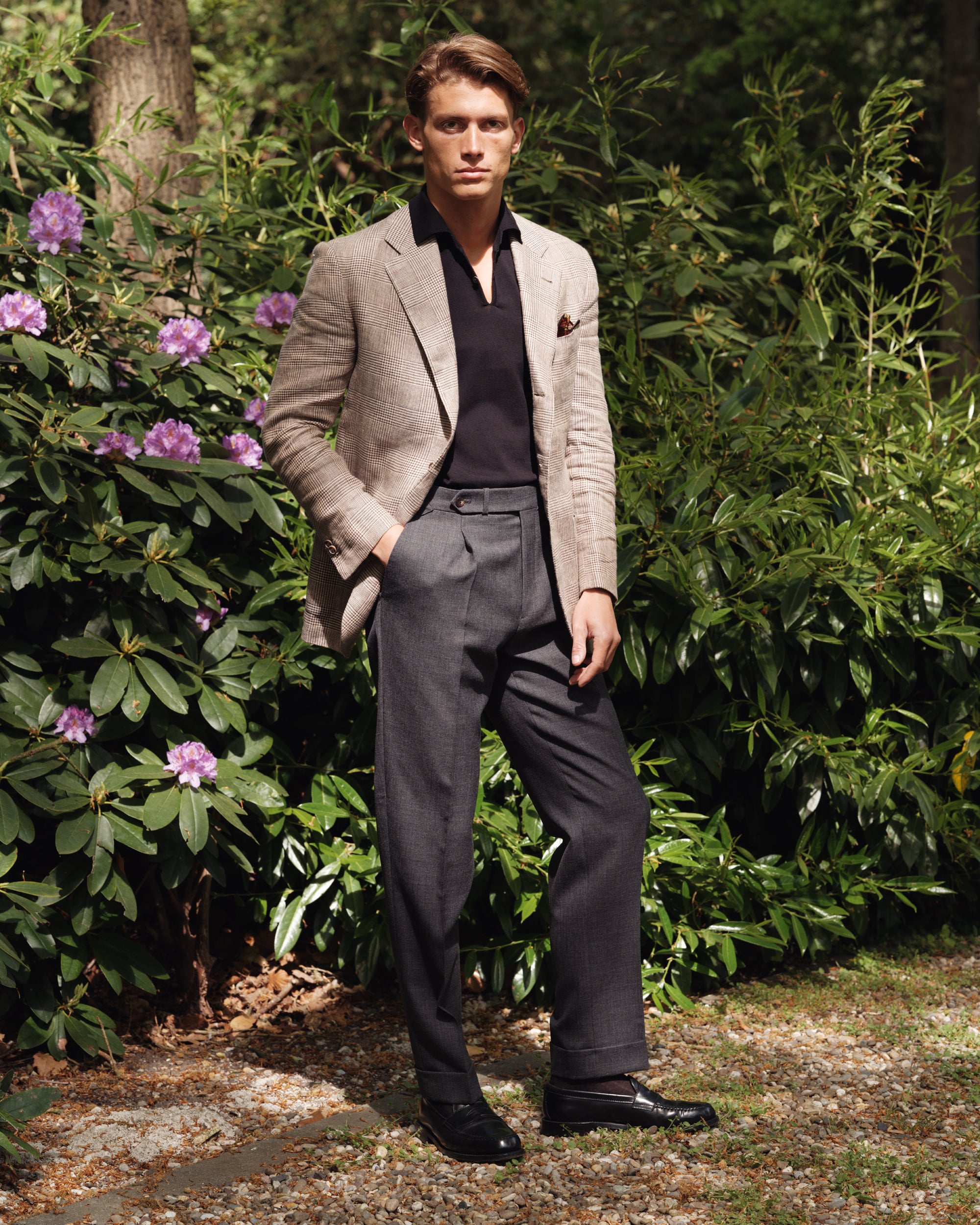Man wearing a brown linen Prince of Wales check jacket, black knit polo shirt, and high-waisted grey wool trousers with black loafers — spring-summer menswear look by Besnard, photographed in a garden setting.