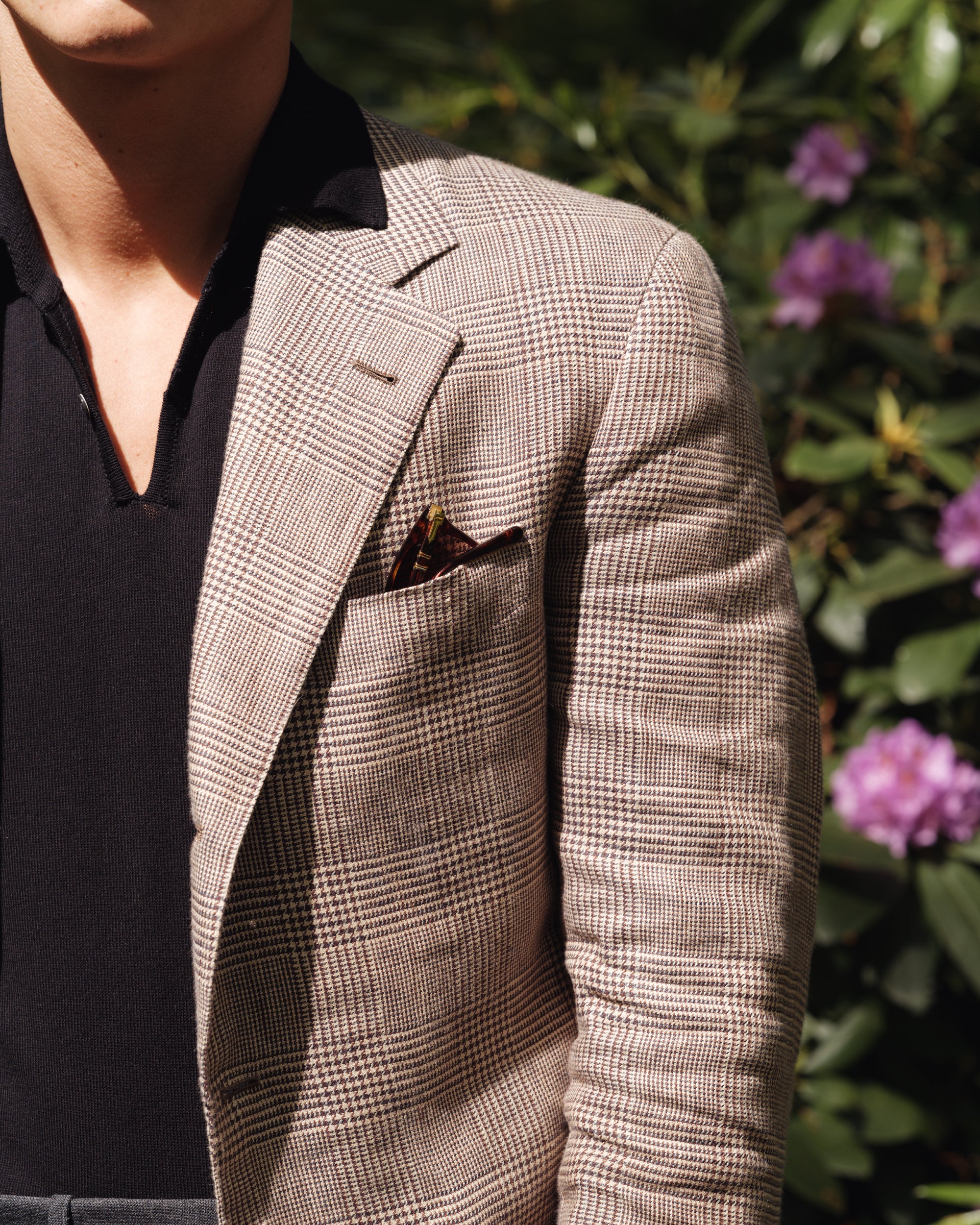 Close-up of a man wearing a brown linen Prince of Wales check jacket with a black knit polo shirt and pocket square — spring-summer menswear look by Besnard.