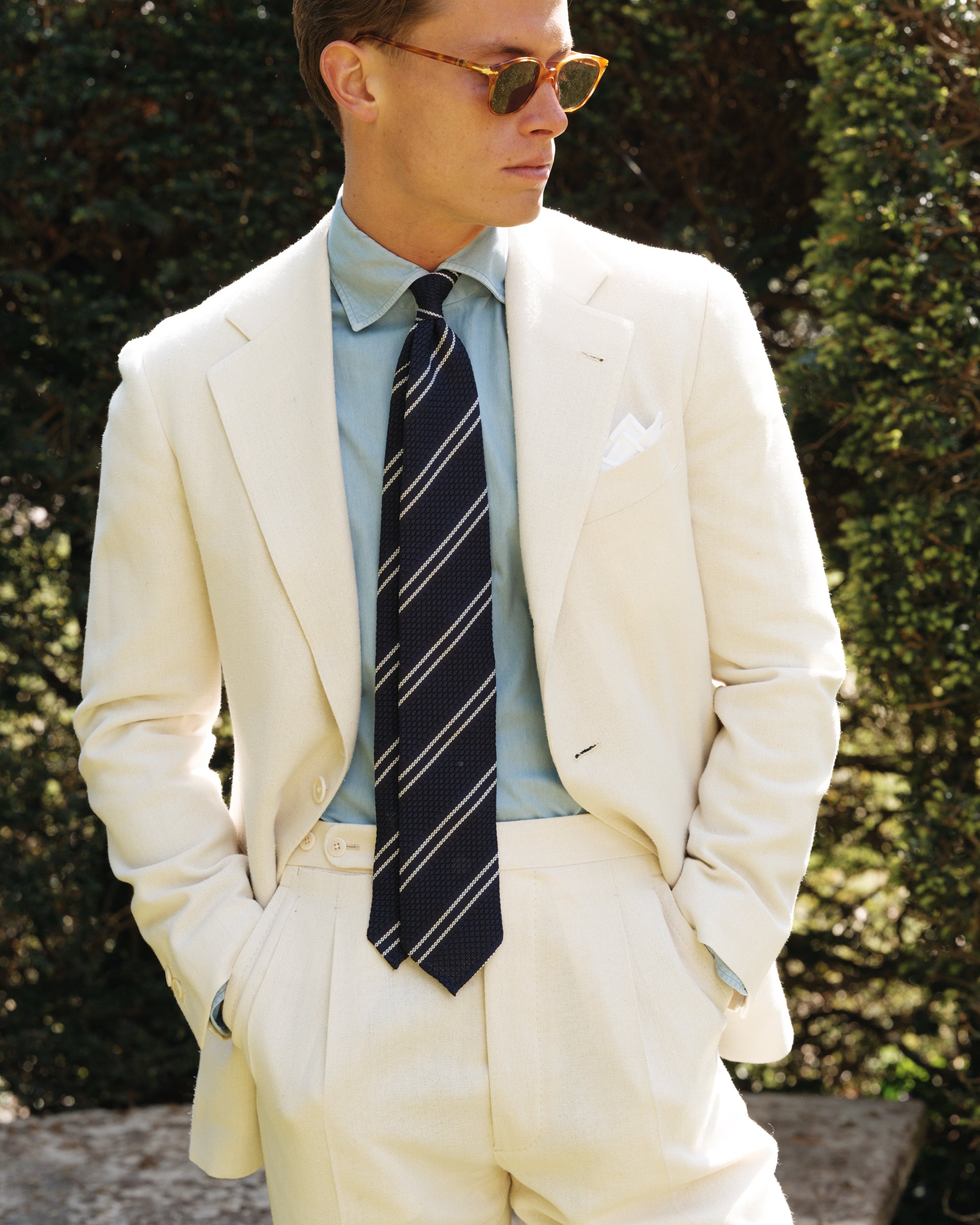 Close-up of a man wearing a cream silk suit, bleached denim shirt, navy striped grenadine tie, and white pocket square — refined spring-summer tailoring by Besnard, photographed in a sunlit garden.