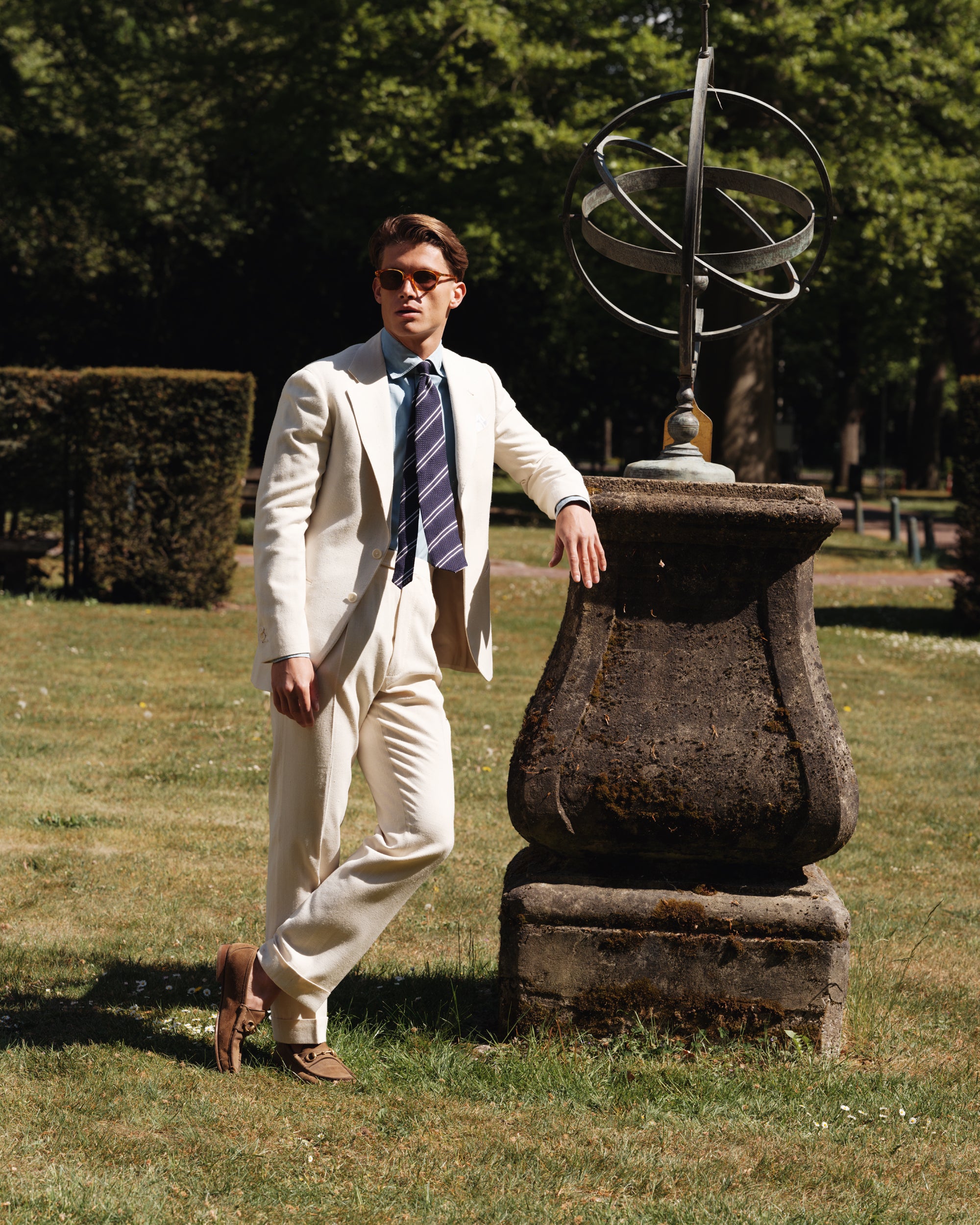 Man wearing a cream silk suit, bleached denim shirt, navy striped grenadine tie, and brown suede Gucci horsebit loafers — elegant spring-summer tailoring by Besnard, photographed in a formal garden beside a sundial sculpture.