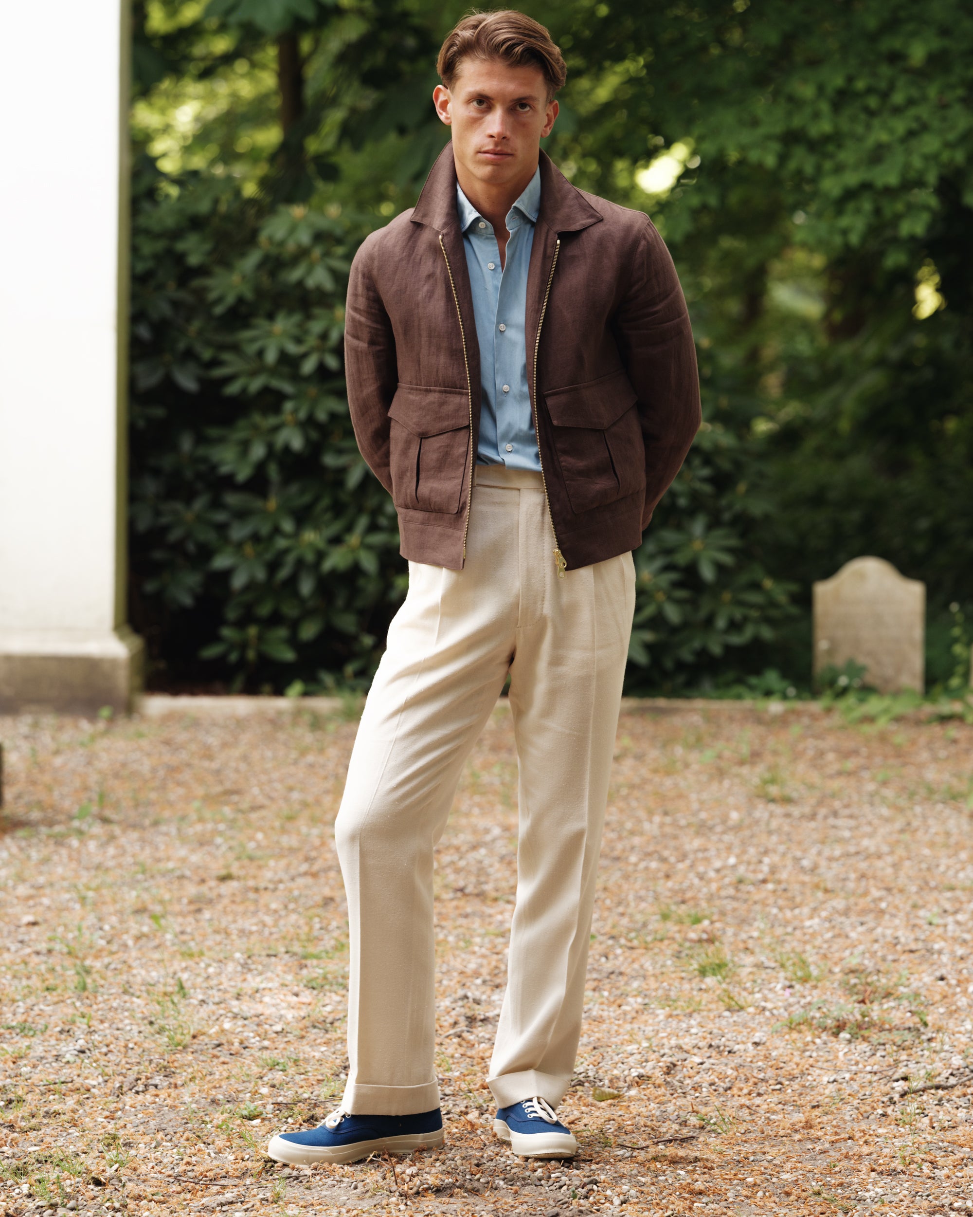 Man wearing a brown linen flight jacket, light blue denim shirt, cream silk trousers, and navy canvas sneakers — casual spring-summer look by Besnard, photographed in a garden setting.