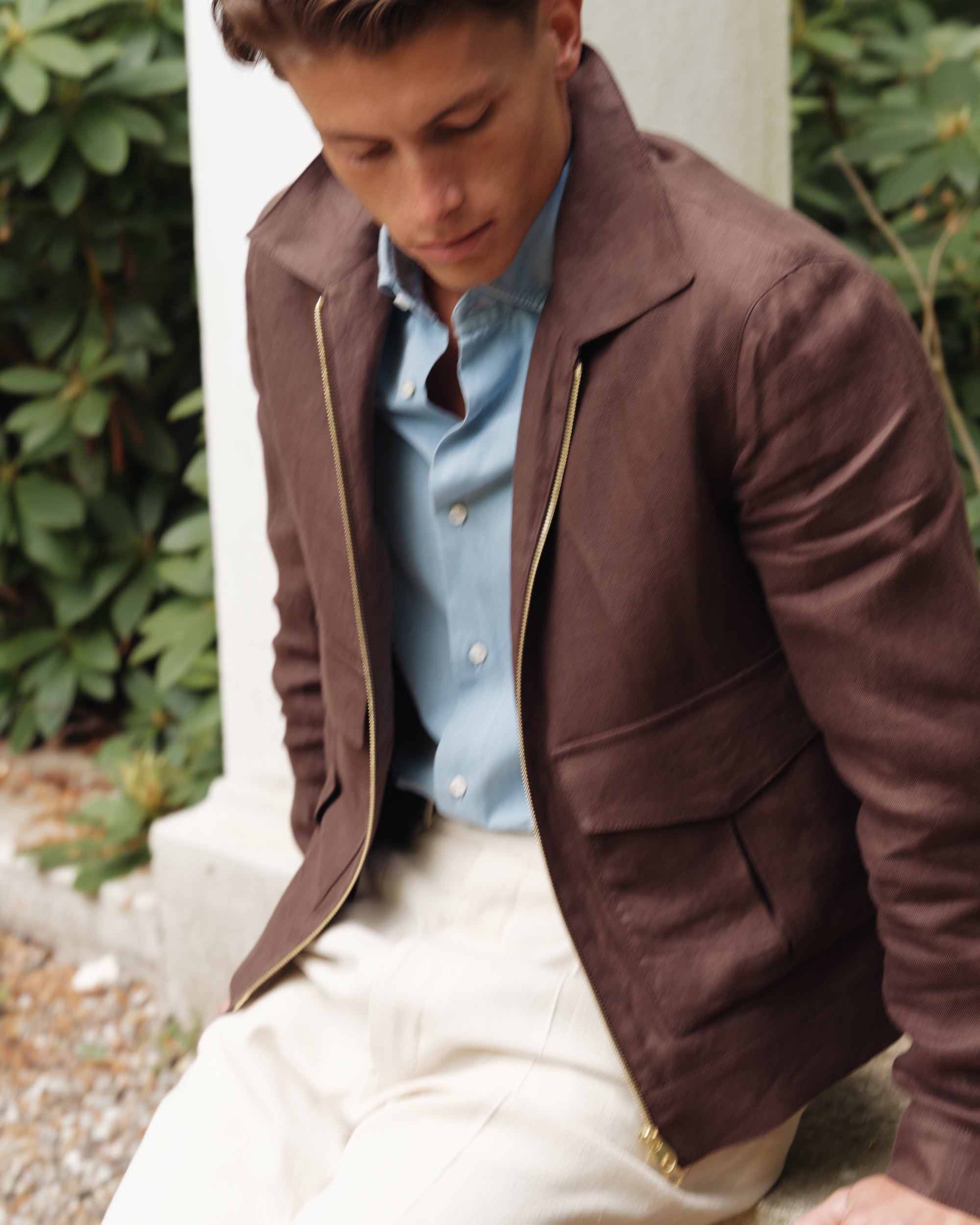 Close-up of a man wearing a brown linen flight jacket, light blue denim shirt, and cream silk trousers — casual spring-summer look by Besnard, photographed in a garden setting.
