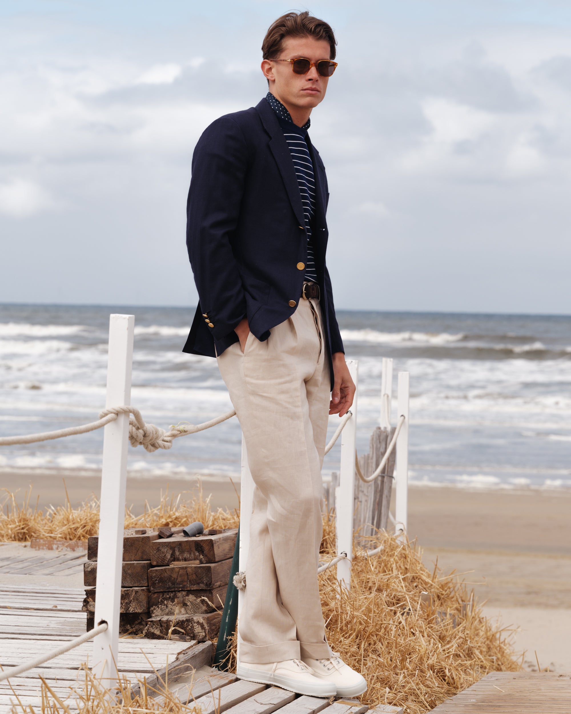 Man wearing a navy wool-linen blazer with gold buttons, navy Breton stripe knit, polka dot neckerchief, stone linen trousers, and white tennis sneakers — nautical-inspired spring-summer look by Besnard, photographed on a coastal boardwalk.