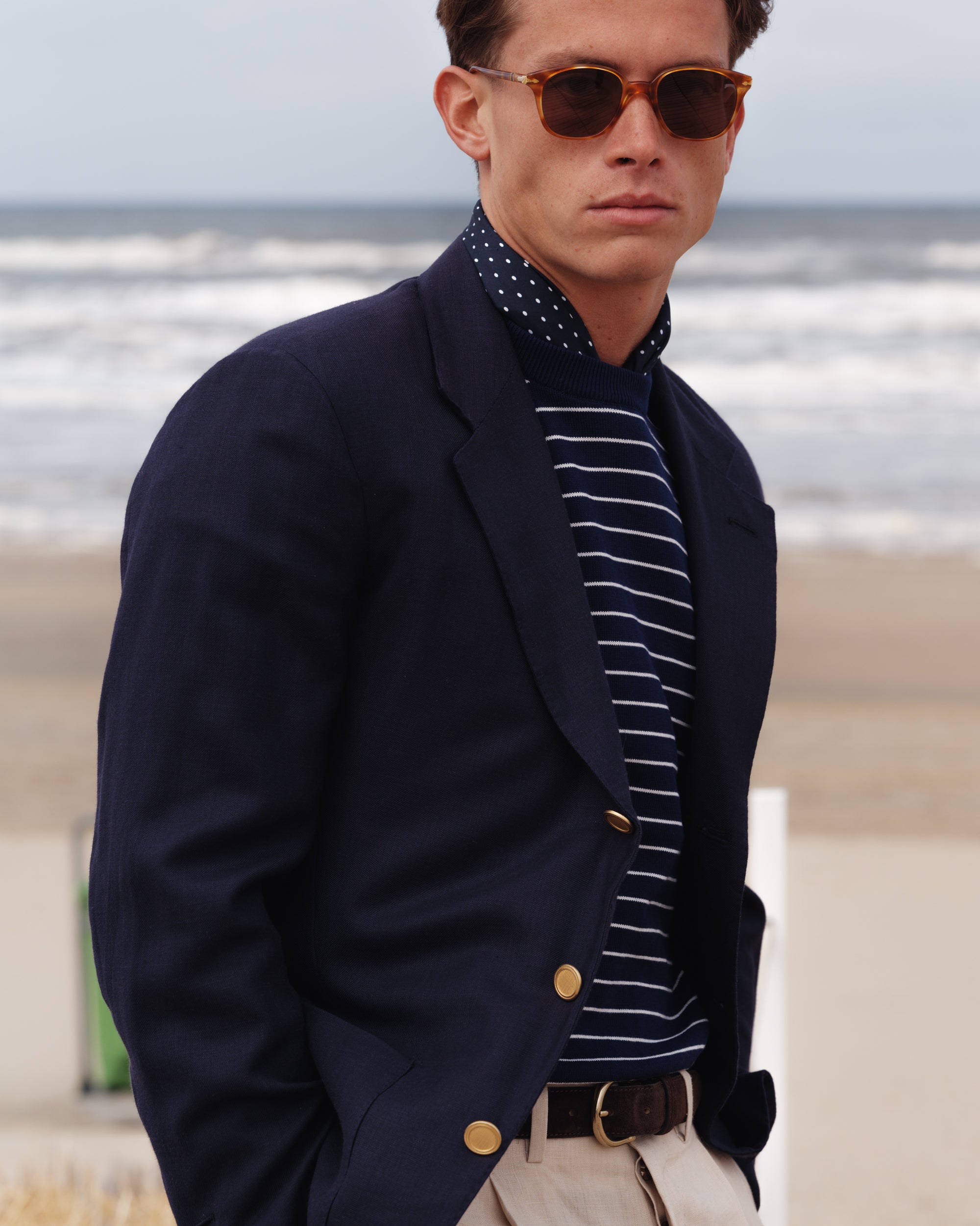 Close-up of a man wearing a navy wool-linen blazer with gold buttons, navy Breton stripe knit, polka dot neckerchief, and stone linen trousers — nautical-inspired spring-summer look by Besnard, photographed on the beach.