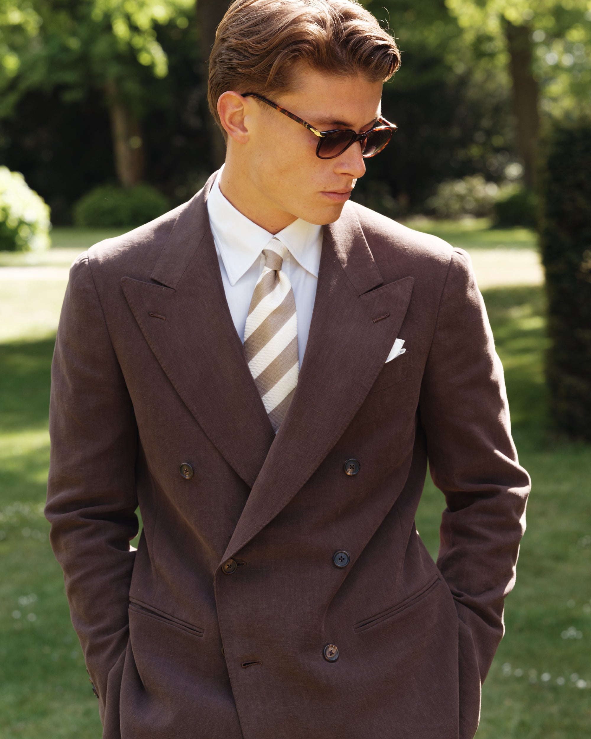 Close-up of a man wearing a brown double-breasted linen suit with wide peak lapels, white point-collar shirt, beige and white striped silk tie, and white pocket square — classic spring-summer tailoring by Besnard, photographed in a sunlit garden.