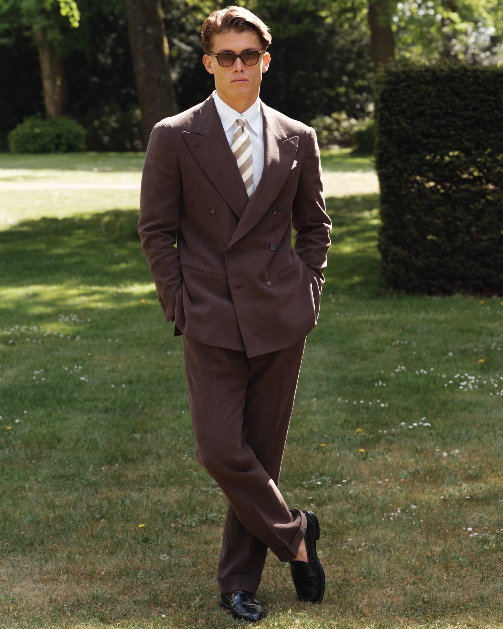 Man wearing a brown double-breasted linen suit with a white point-collar shirt, beige and white striped silk tie, and black cordovan penny loafers — classic spring-summer tailoring by Besnard, photographed in a sun-lit garden.