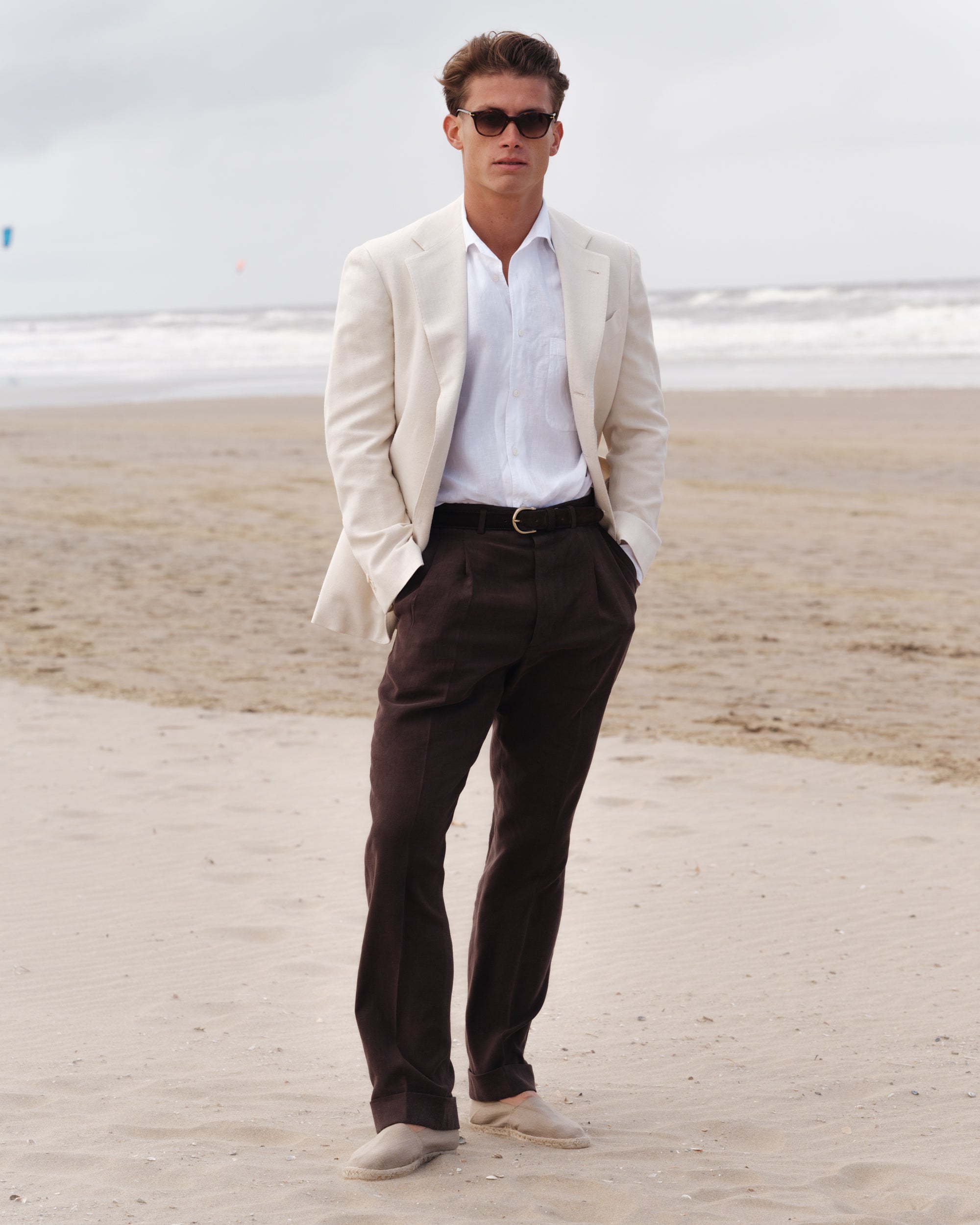 Man wearing a cream linen jacket, white linen shirt with a one-piece collar, dark brown linen trousers, and beige espadrilles — relaxed spring-summer menswear look by Besnard, photographed on a breezy beach.