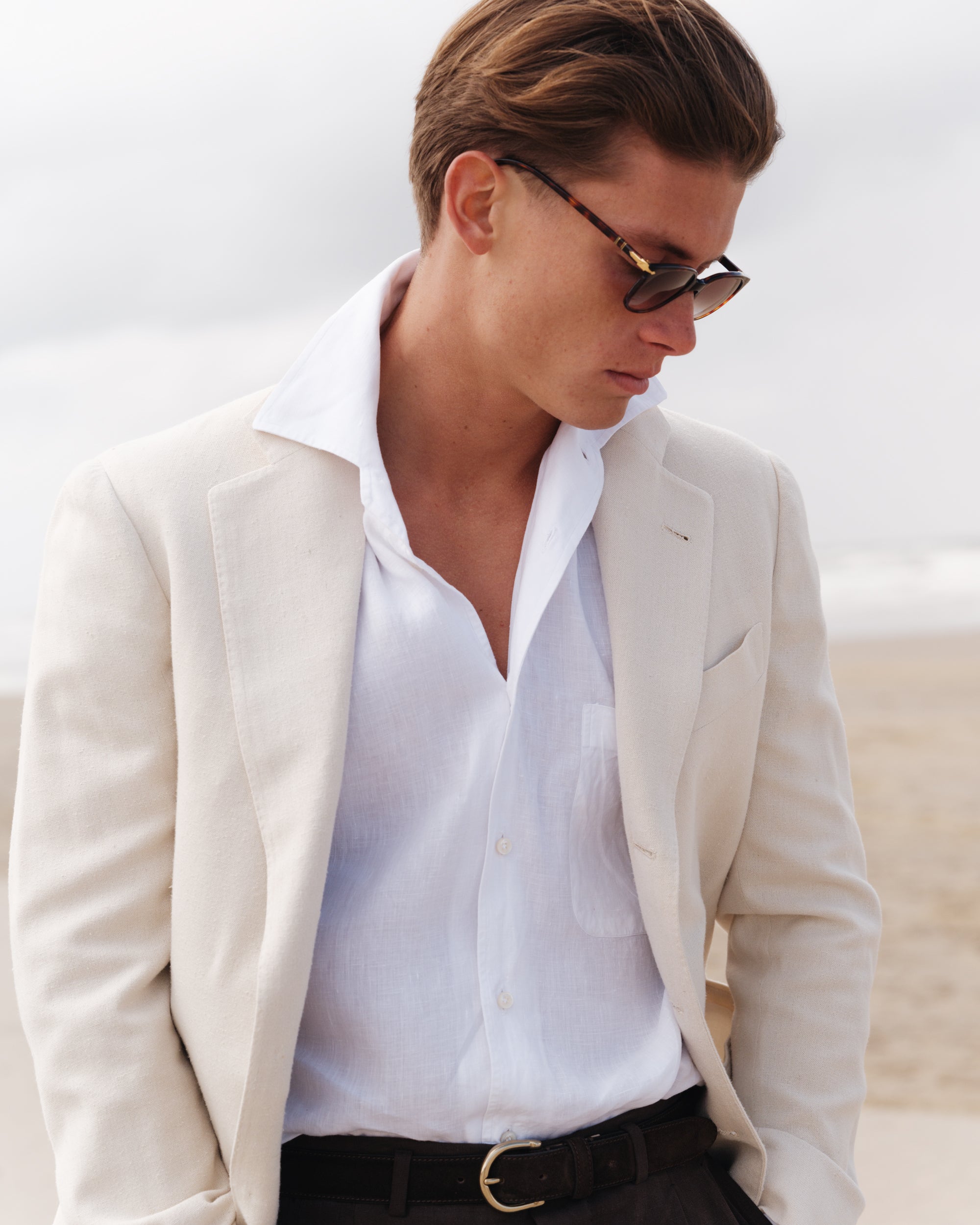 Close-up of a man wearing a cream linen jacket, white linen shirt with a one-piece collar, and dark brown trousers with a brass buckle belt — relaxed spring-summer look by Besnard, photographed on a windy beach.