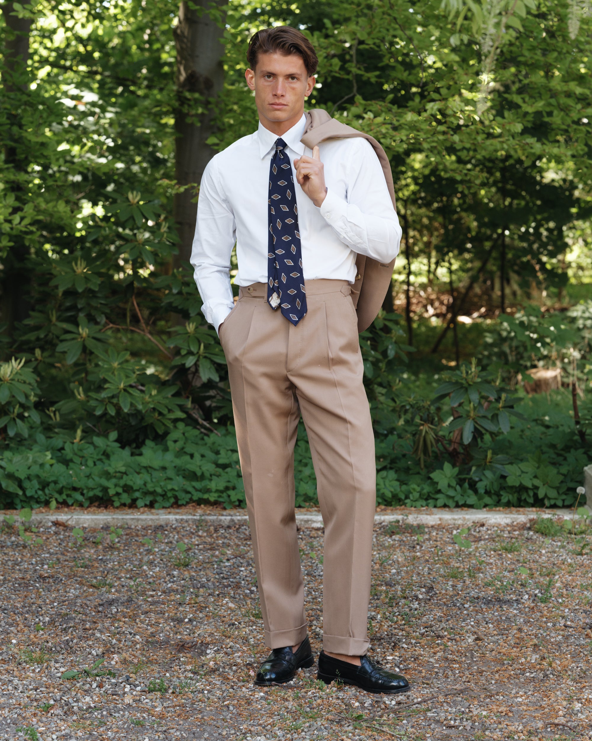 Man wearing tan wool gabardine trousers with a white point collar shirt, navy patterned silk tie, and black cordovan penny loafers, holding the matching jacket over his shoulder — classic spring-summer tailoring by Besnard, photographed in a garden setting.