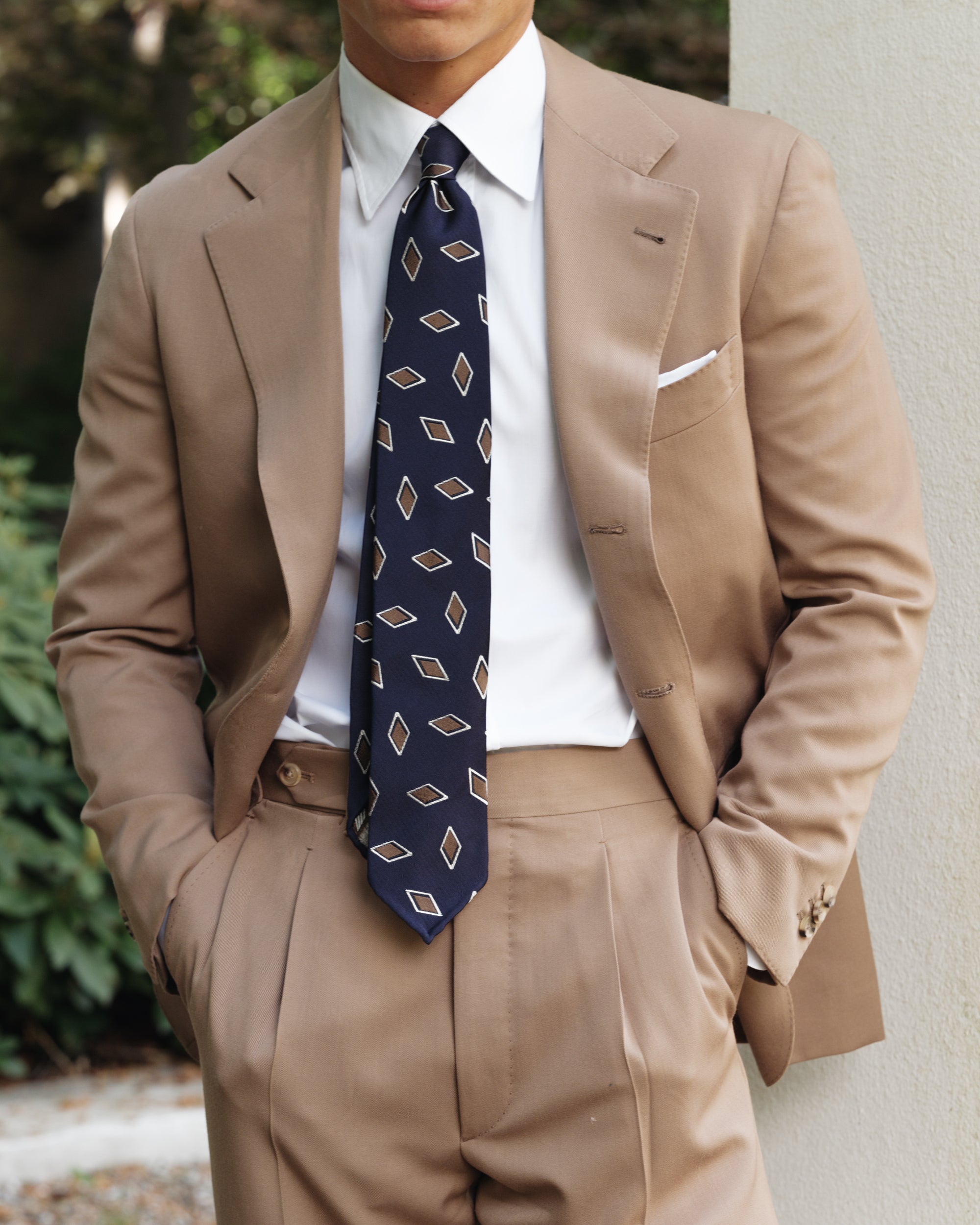 Close-up of a man wearing a tan wool gabardine suit with a white point collar shirt, navy patterned silk tie, and white pocket square — elegant spring-summer tailoring by Besnard.