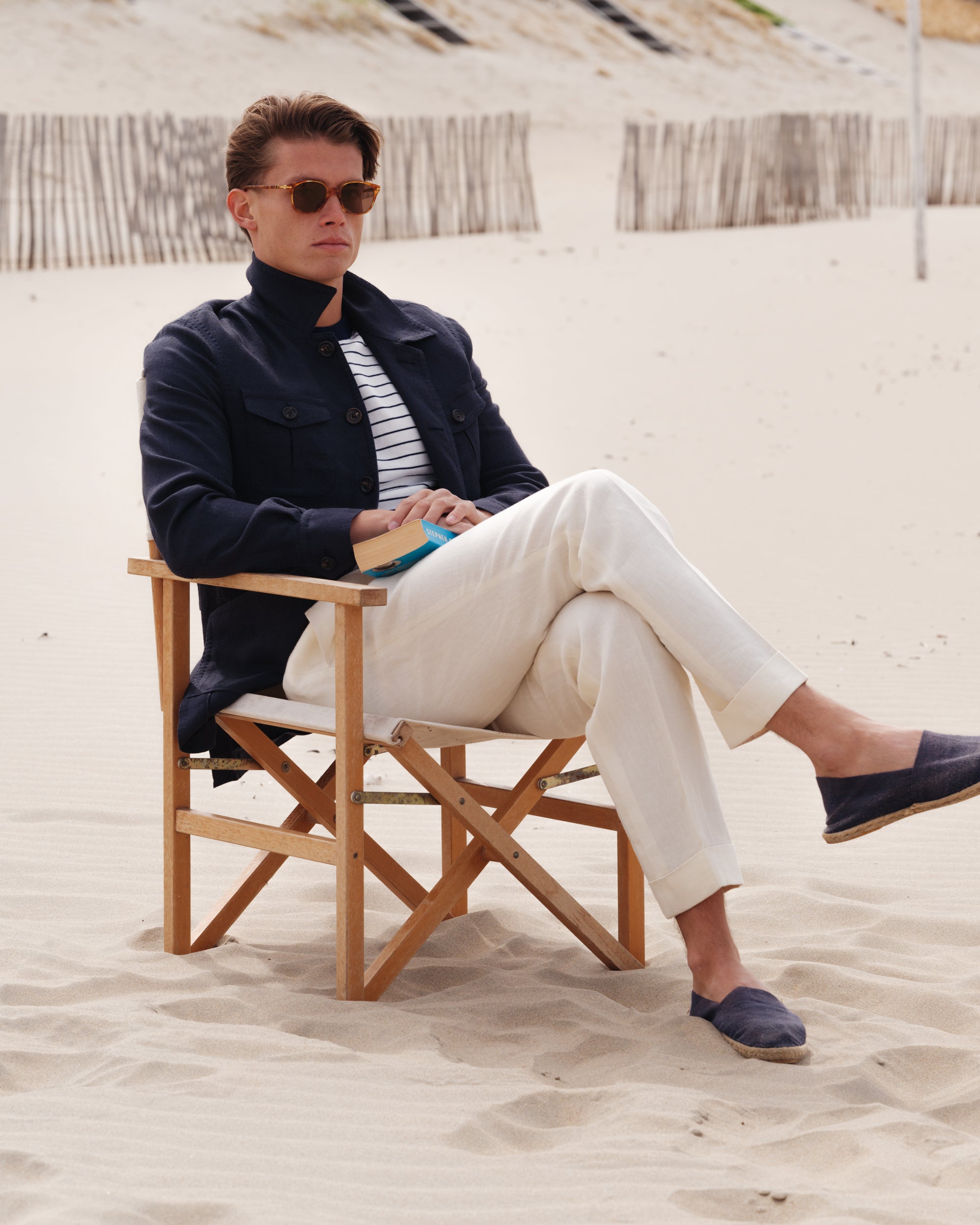 Man seated on a wooden chair in the sand, wearing a navy safari jacket over a white Breton stripe T-shirt, off-white linen trousers, and navy espadrilles — relaxed spring-summer menswear look by Besnard, photographed on a windswept beach.