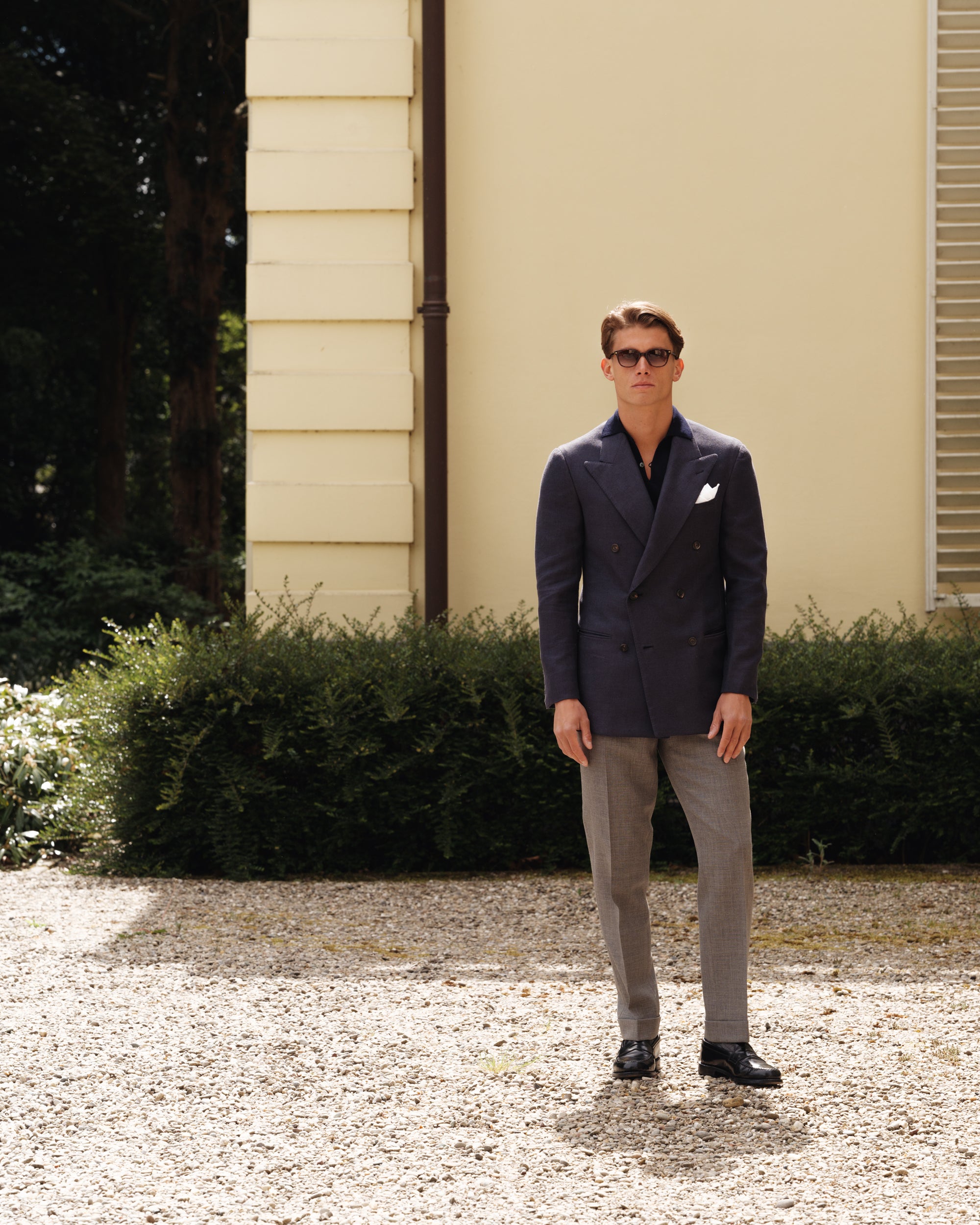 Man wearing a navy double-breasted jacket, navy knit polo, grey tropical wool trousers, and black cordovan penny loafers with a white pocket square — refined spring-summer menswear look by Besnard, photographed in a sunlit villa courtyard.