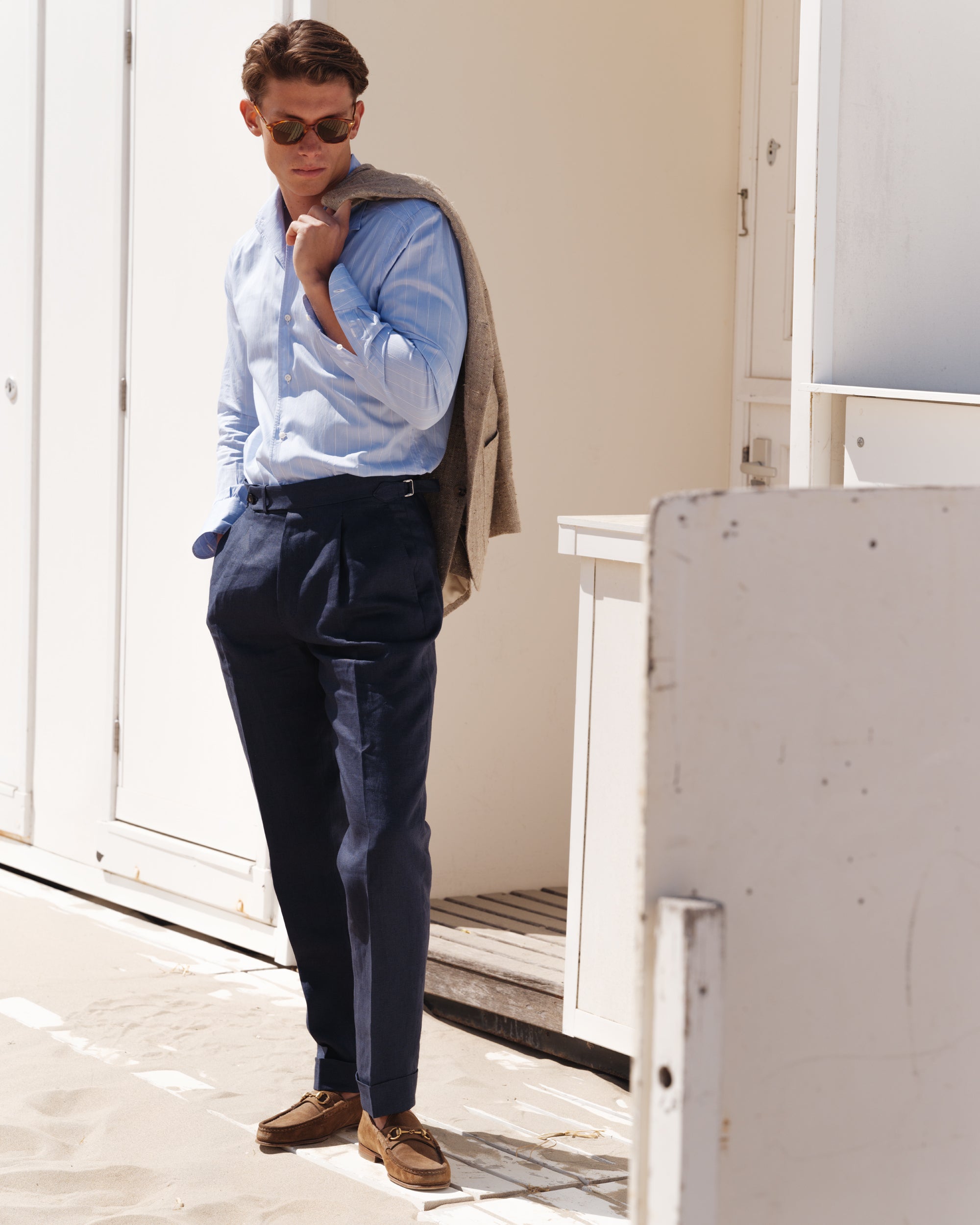 Man wearing a light blue striped shirt and navy high-waisted linen trousers, with a beige silk-linen jacket draped over his shoulder and brown suede Gucci horsebit loafers — elegant spring-summer menswear look by Besnard, photographed on a sandy boardwalk.