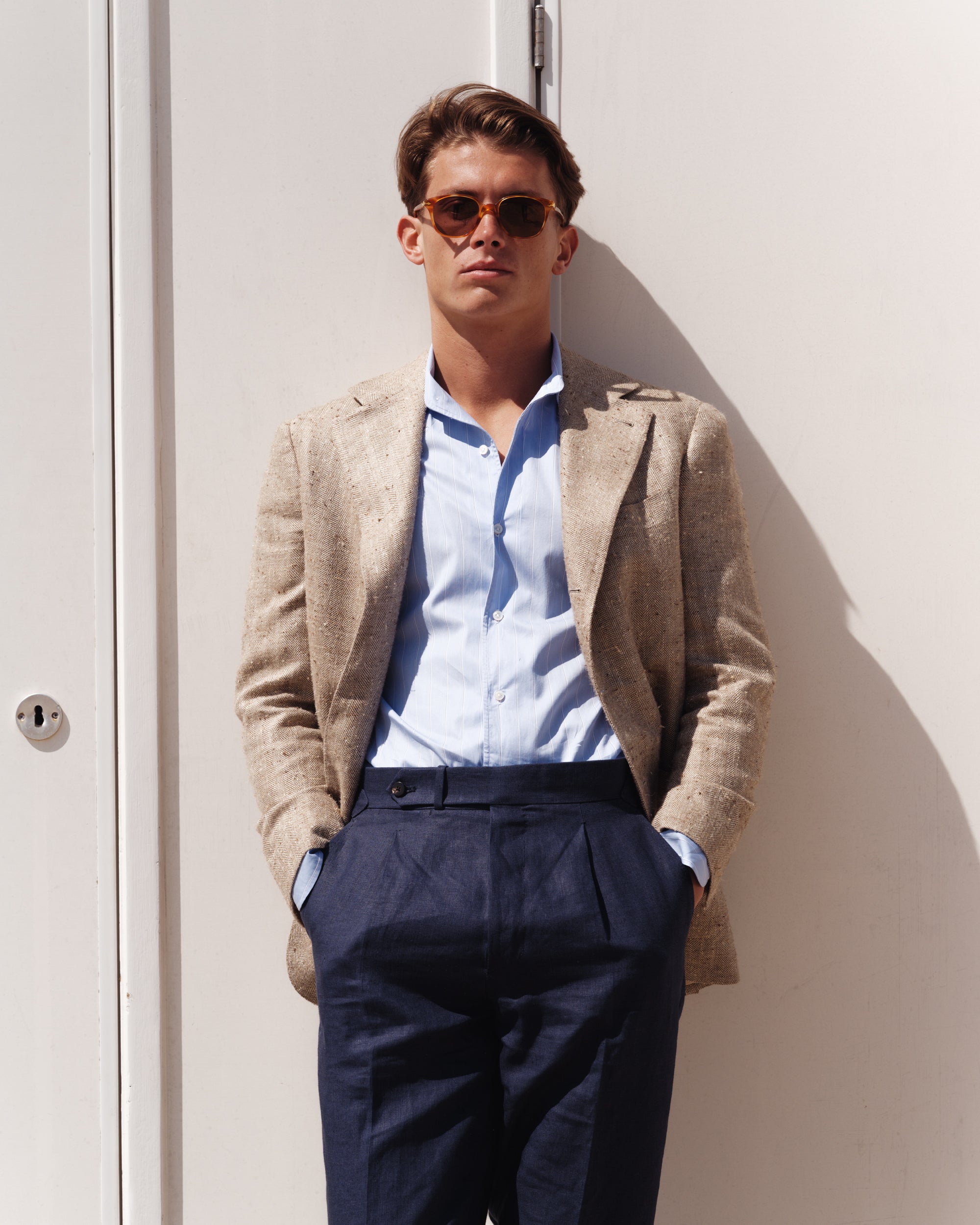 Man wearing a beige silk-linen jacket, light blue striped shirt, and navy high-waisted linen trousers — tailored spring-summer menswear look by Besnard, photographed in bright sunlight against a white beach hut.